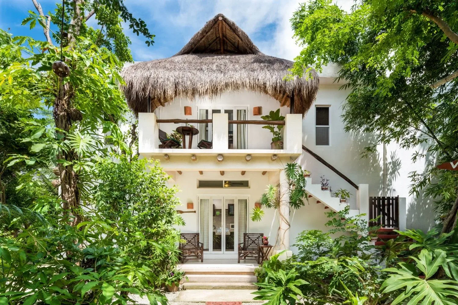 Edificio blanco de hotel con techo de paja entre plantas tropicales.