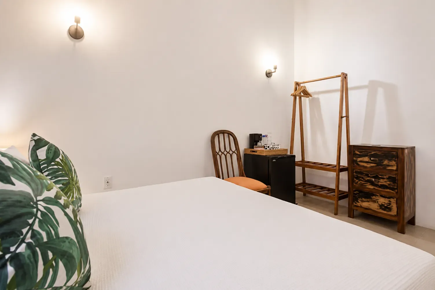 Dormitorio con una cama blanca, una silla de mimbre y una planta en maceta.