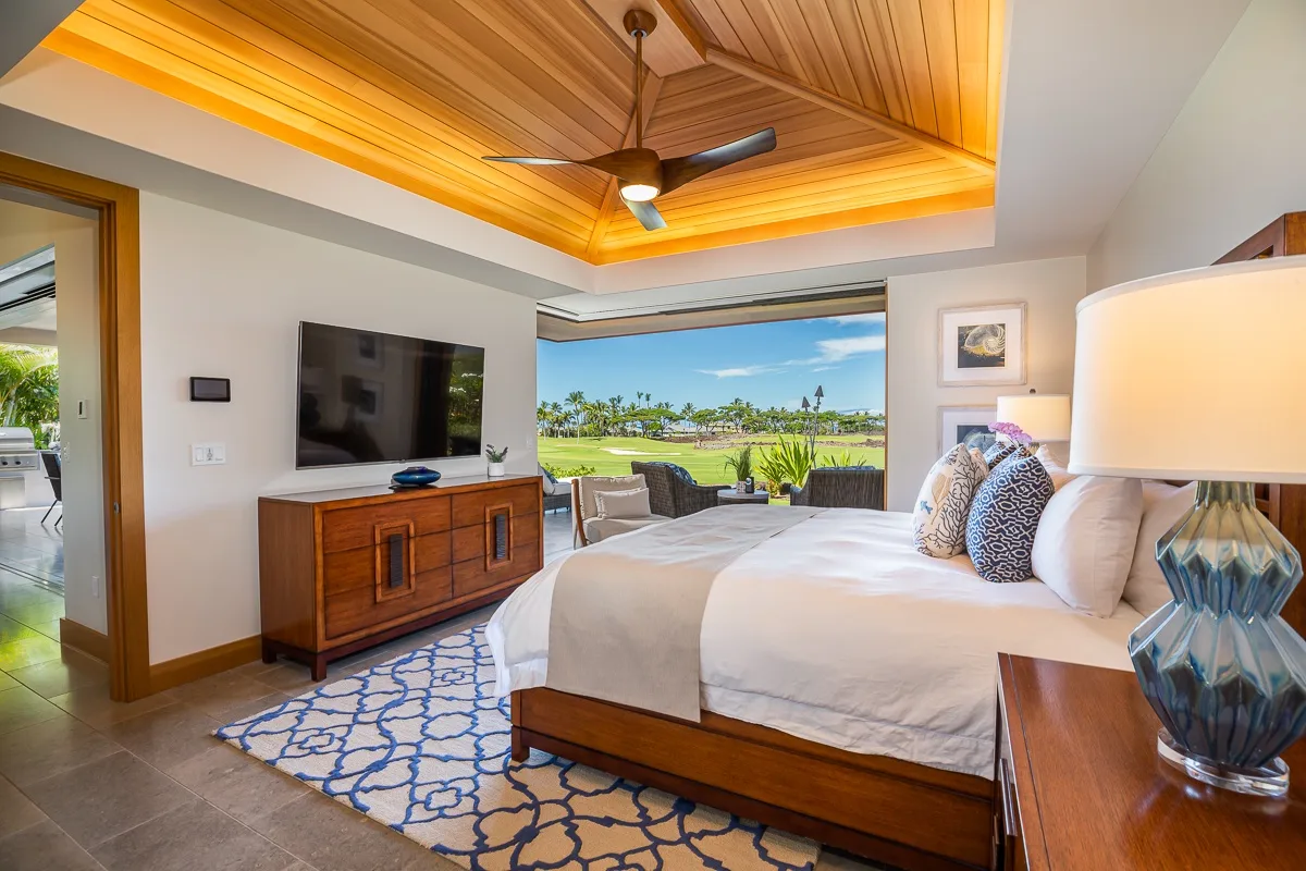 A bedroom with a large bed, wooden ceiling, and a view of greenery through the windows.