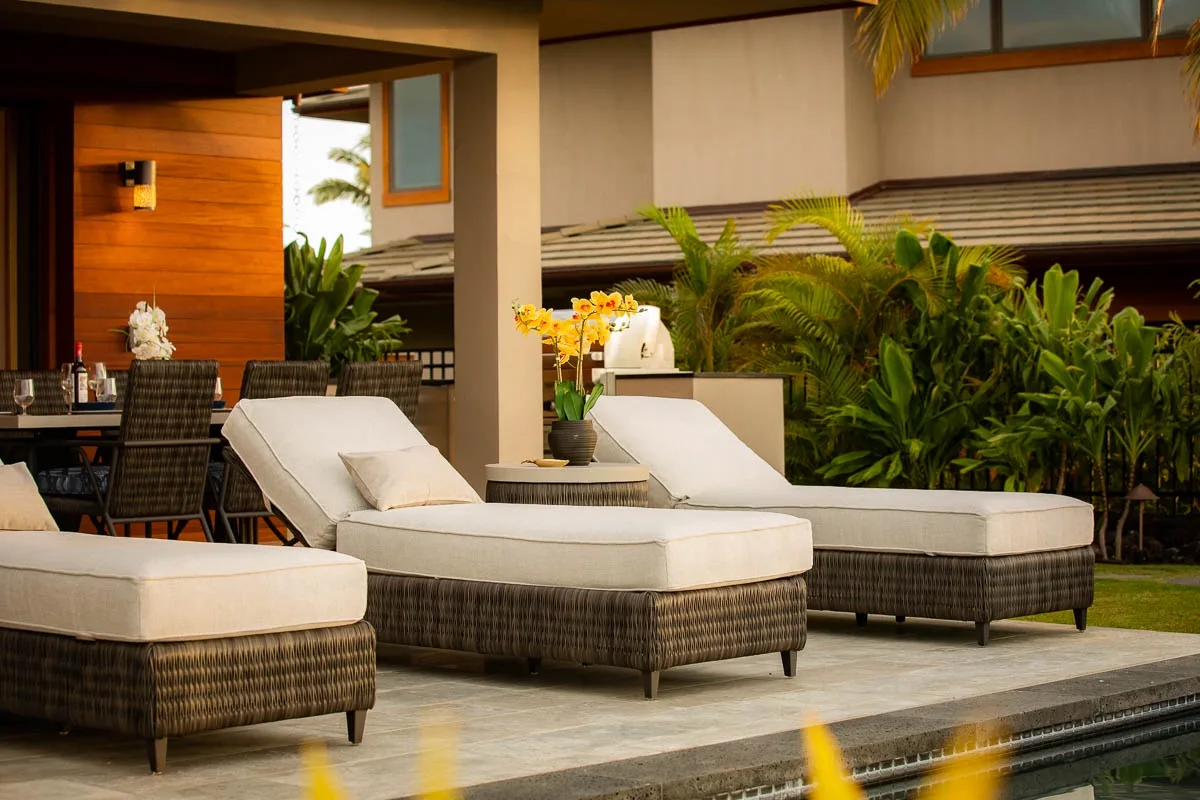 An outdoor patio has cushioned lounge seating beside a pool and leafy plants.