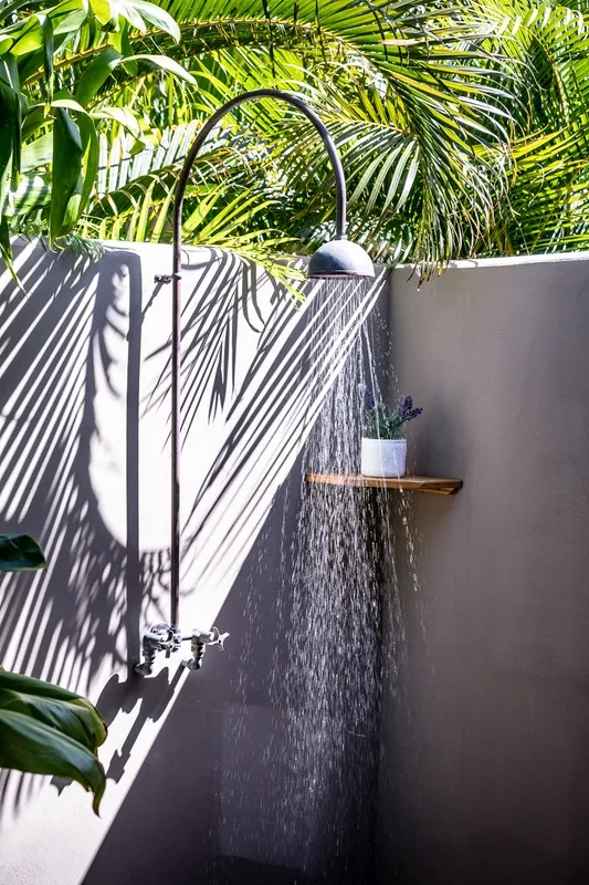 An outdoor shower stands beside a small wall shelf amid tropical foliage.