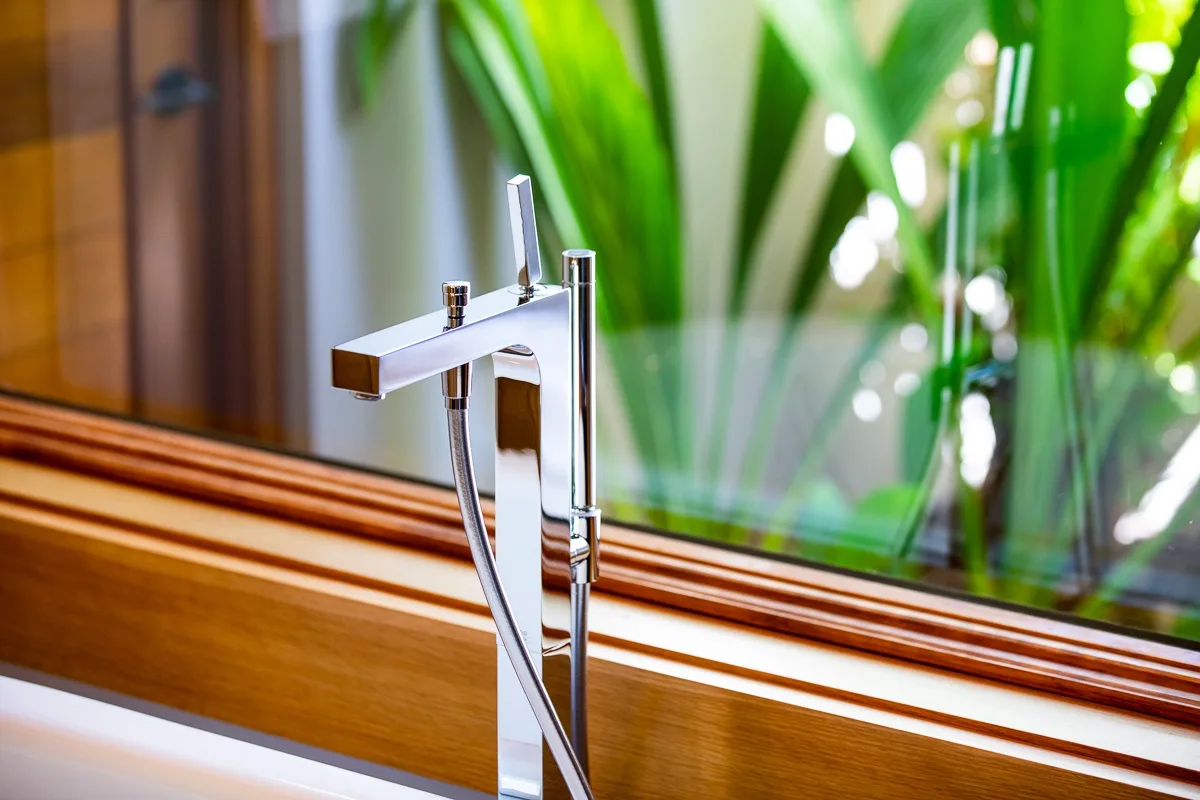 A bathtub faucet stands beside a wooden-framed window overlooking green plants.
