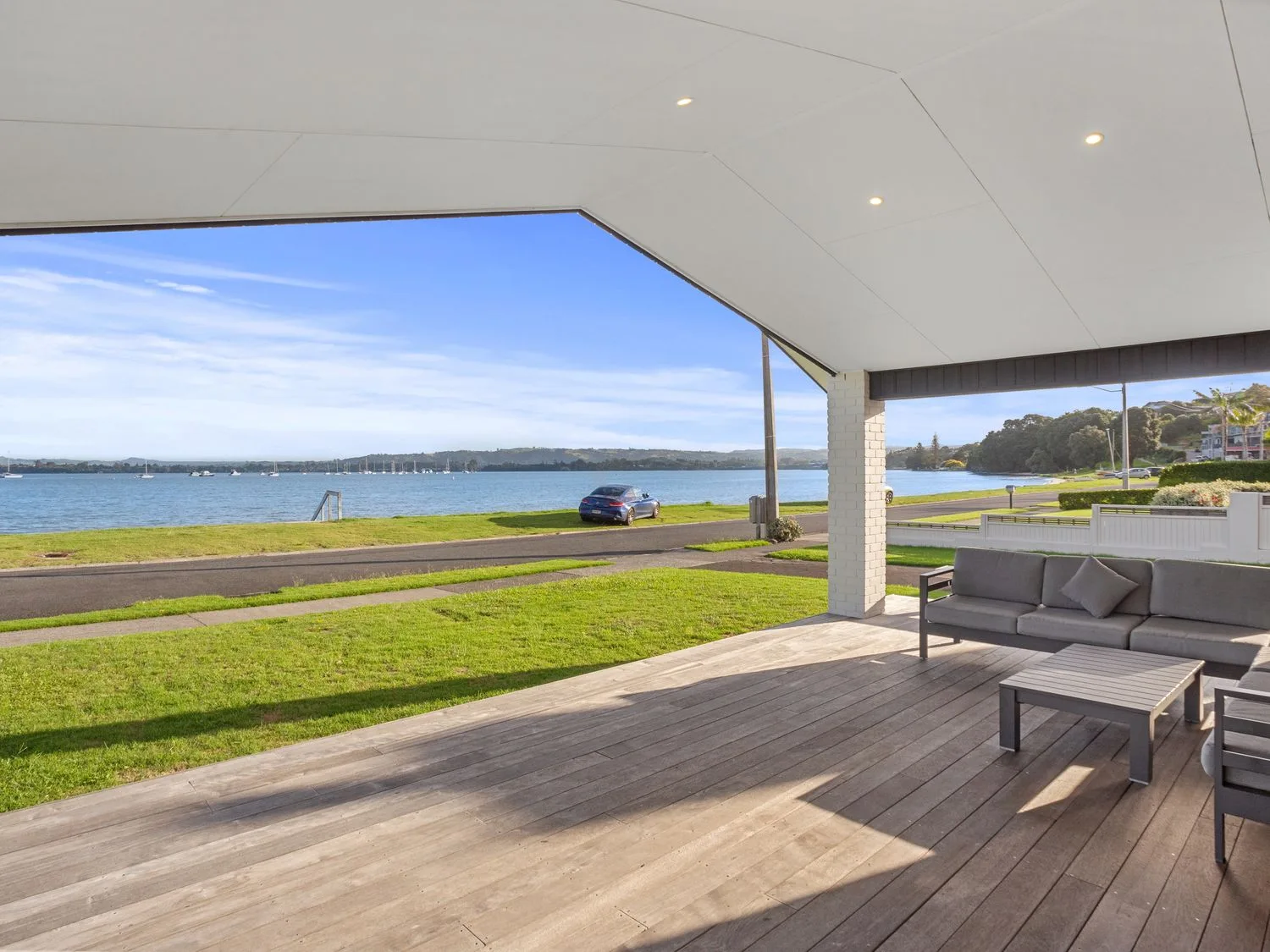 Una terraza cubierta de madera con asientos de exterior orientados hacia una carretera y el agua con barcos en Waterfront Oasis Omokoroa Holiday Home, en Omokoroa