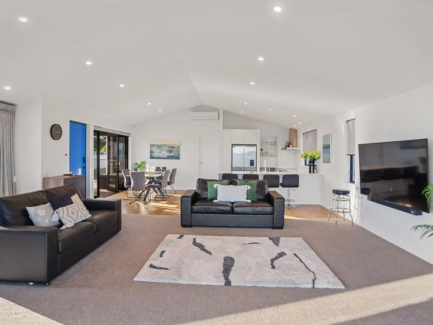 Una sala de estar con dos sofás de cuero negro, una alfombra y un televisor montado en la pared, con una zona de comedor y una cocina al fondo, en Waterfront Oasis - Omokoroa Holiday Home, en Omokoroa
