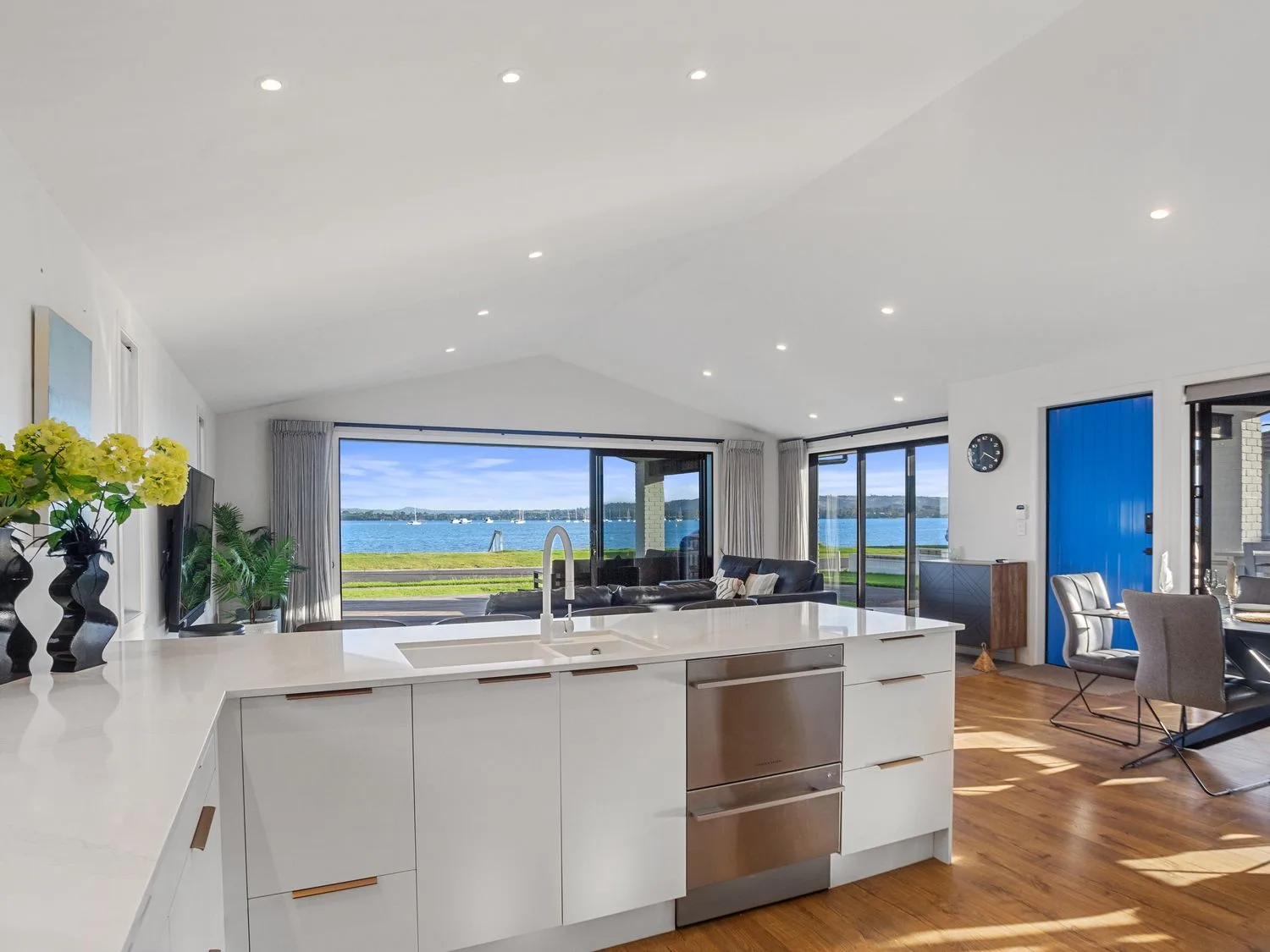 Una cocina con armarios blancos y una isla que da a una sala de estar y una zona de comedor con una puerta azul y vistas al agua en Waterfront Oasis Omokoroa Holiday Home, en Omokoroa