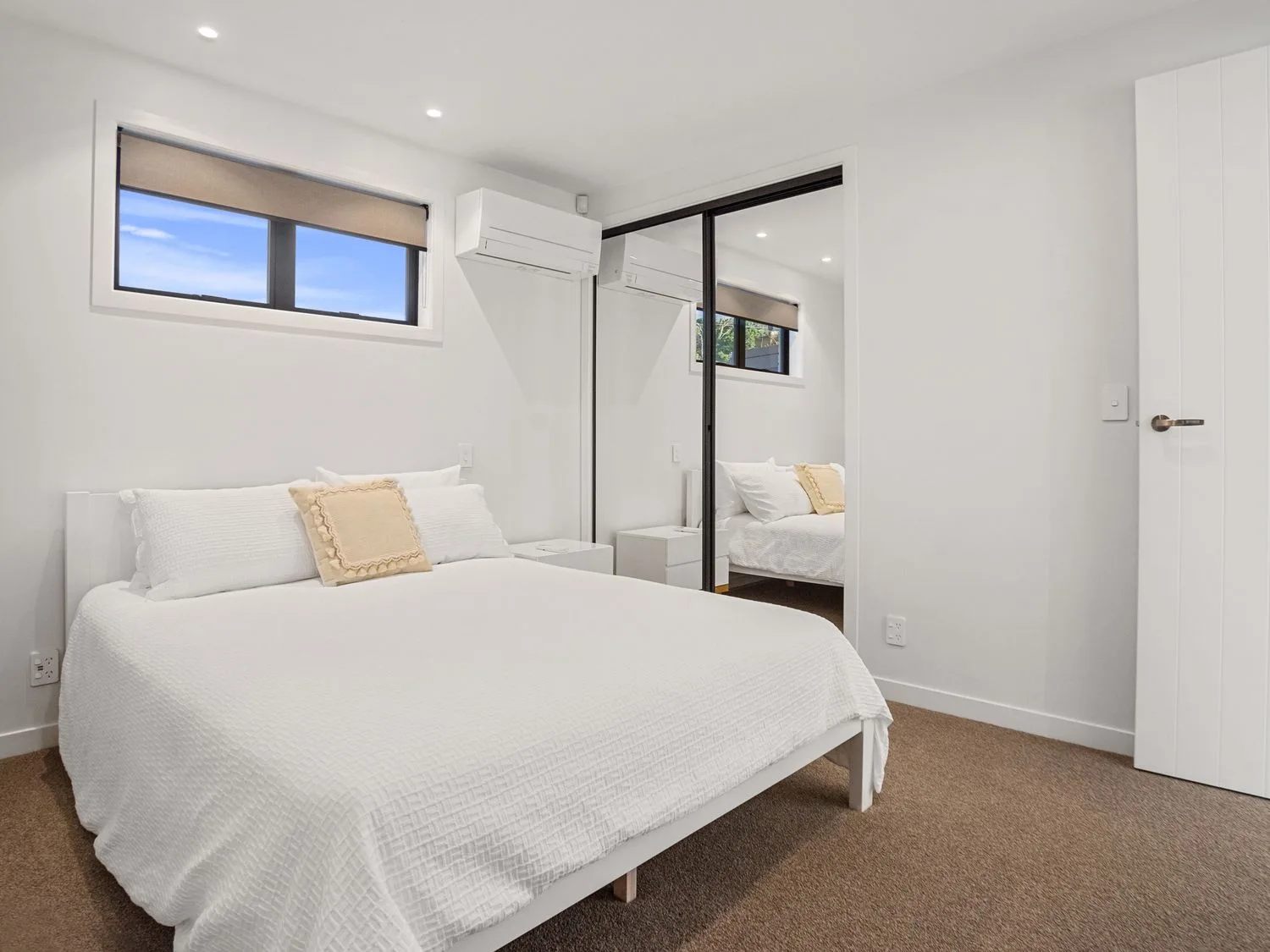 Un dormitorio con una cama con ropa de cama blanca, un cojín pequeño, mesas de noche, un armario con espejo y una ventana en Waterfront Oasis - Omokoroa Holiday Home, en Omokoroa