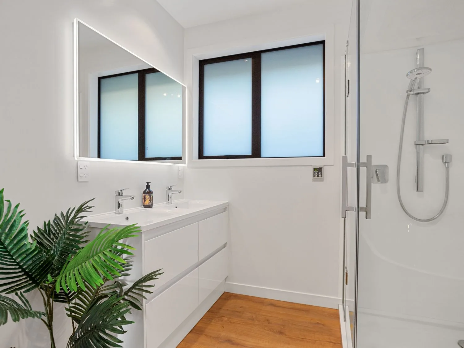 Un baño luminoso con mueble de lavabo, espejo, ducha y una planta frondosa.