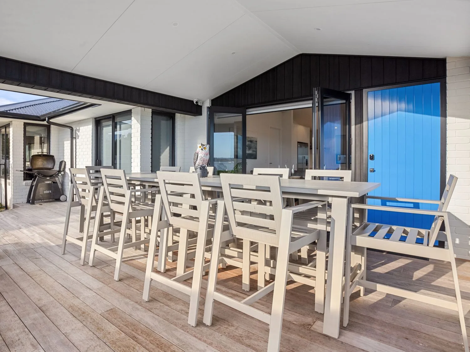 Una zona de comedor al aire libre con una mesa larga y sillas junto a una puerta azul y una barbacoa en Waterfront Oasis - Omokoroa Holiday Home, en Omokoroa