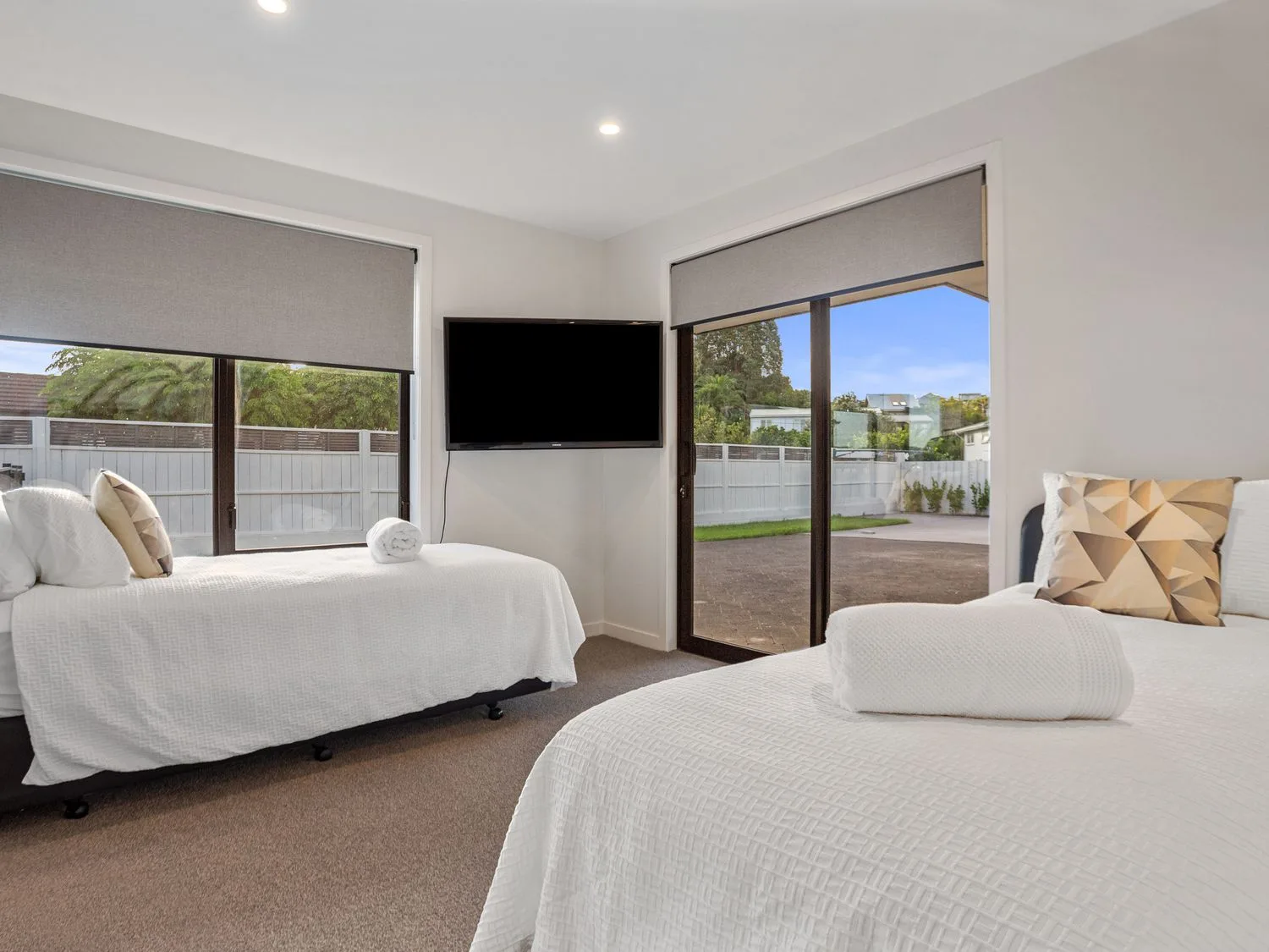 Un dormitorio con dos camas con ropa de cama blanca, un televisor montado en la pared y ventanas grandes en Waterfront Oasis - Omokoroa Holiday Home, en Omokoroa