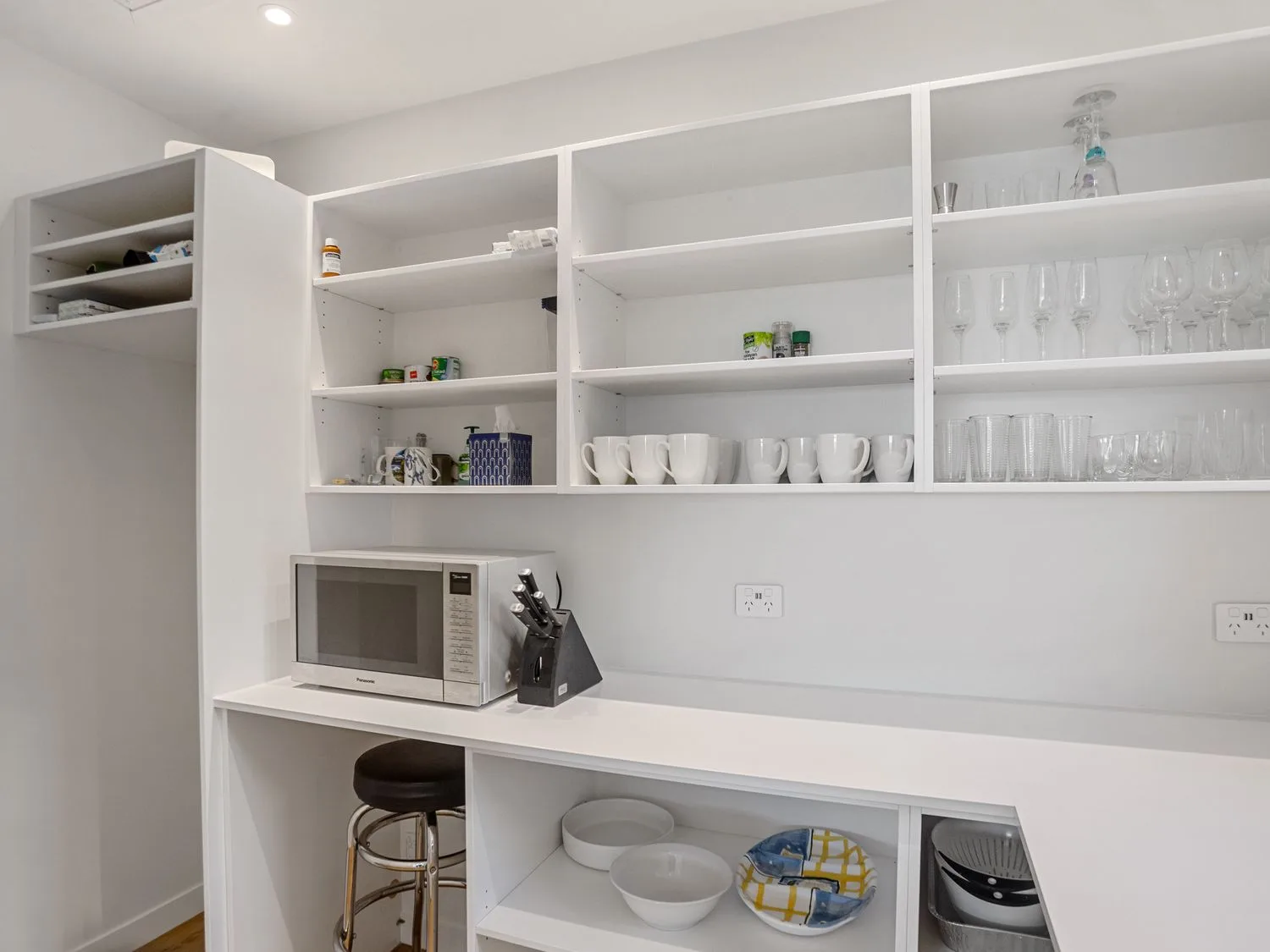 Una cocina pequeña con estantes blancos que contienen tazas, vasos, cuencos, un microondas y un bloque para cuchillos, con un taburete debajo, en Waterfront Oasis - Omokoroa Holiday Home, en Omokoroa