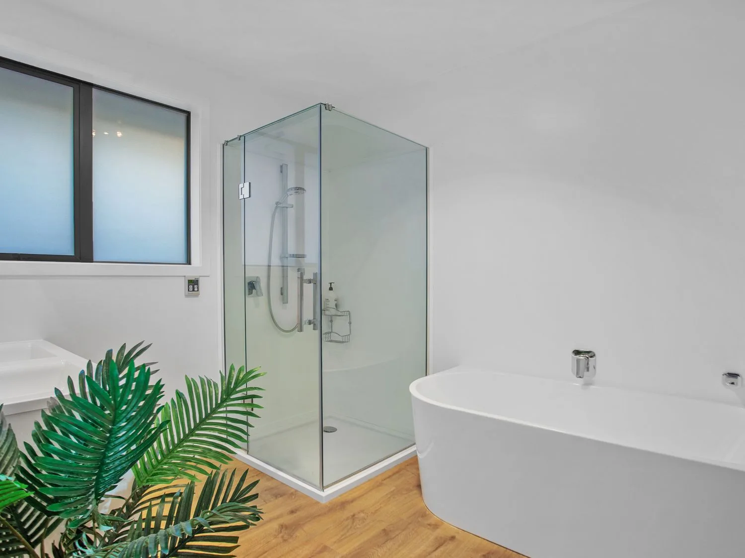Un baño con una ducha con mampara de cristal, una bañera independiente, una ventana y una planta sobre un suelo de madera en Waterfront Oasis - Omokoroa Holiday Home, en Omokoroa