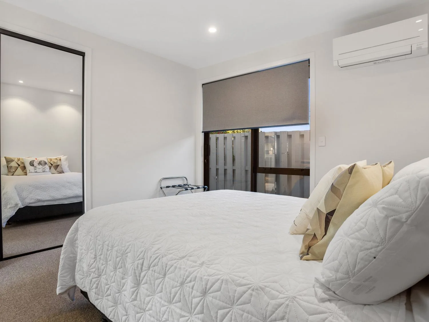 Un dormitorio con una cama cubierta con una colcha blanca y almohadas, una puerta corredera de armario con espejo, una ventana con una persiana enrollable y un aparato de aire acondicionado en Waterfront Oasis - Omokoroa Holiday Home, en Omokoroa