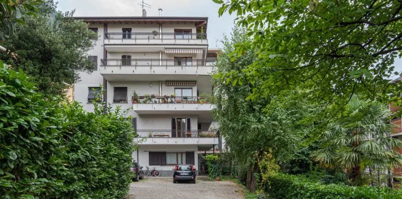 Un edificio residencial de varias plantas se alza tras un jardín frondoso y una entrada.