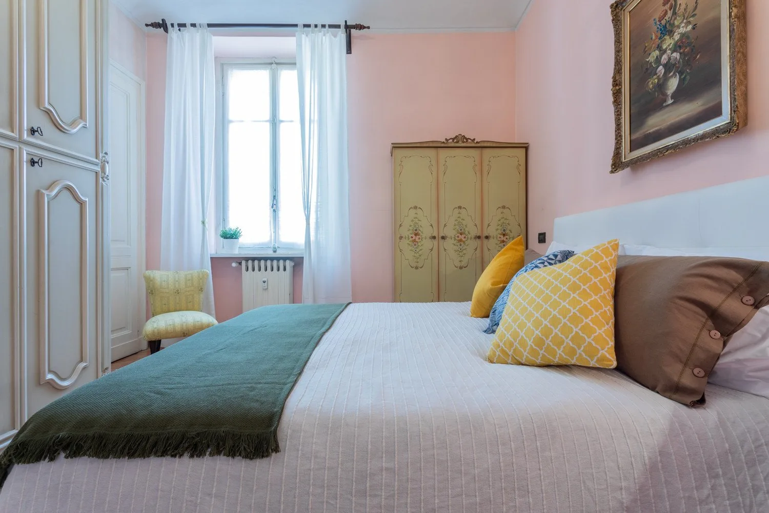 A bedroom with a neatly made bed, colorful pillows, and a tall window.