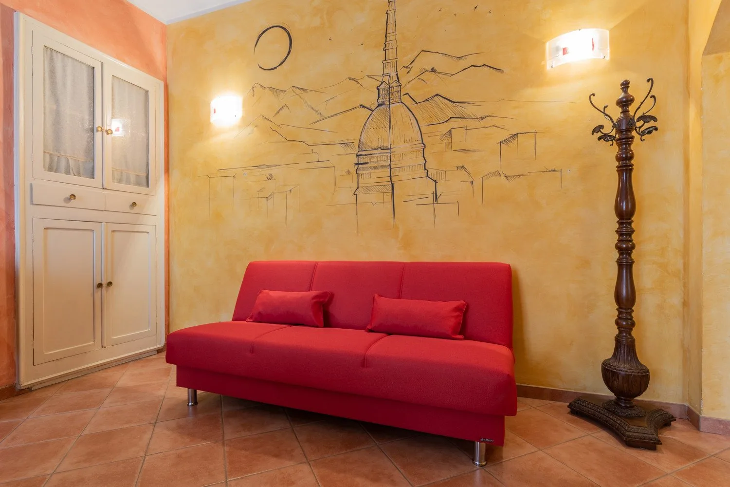 A red seating bench stands against a wall with a decorative cityscape drawing.