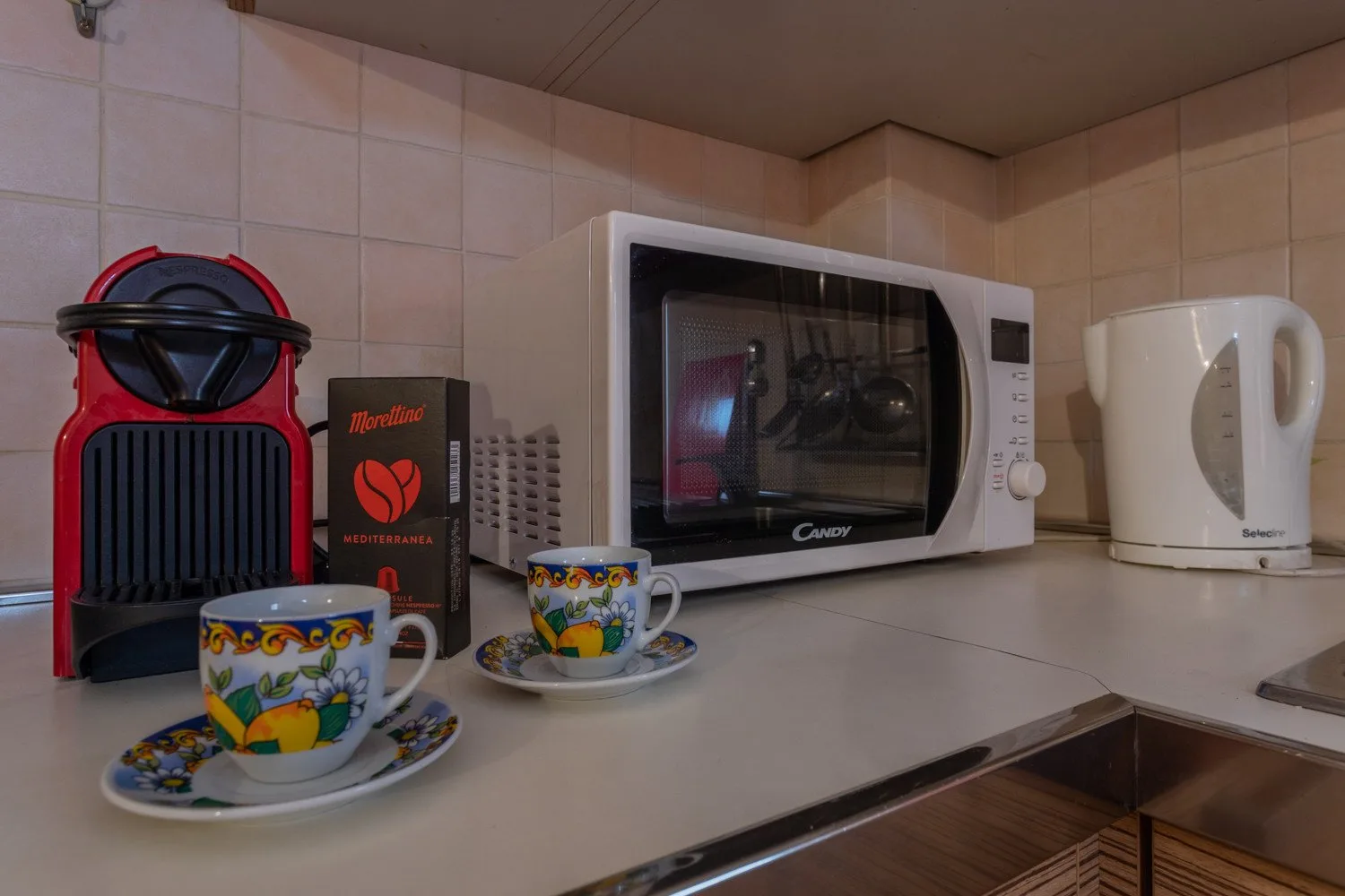 A tiled kitchen counter holds a microwave, kettle, coffee maker, and cups.