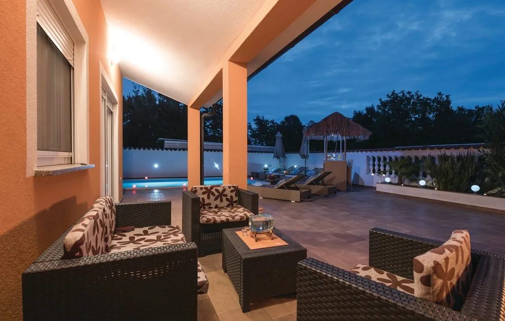 A covered outdoor patio with seating overlooks an illuminated swimming pool at dusk.