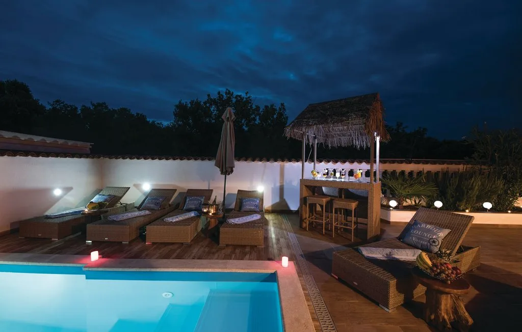 A poolside patio at night has lounge seating and a thatched bar.