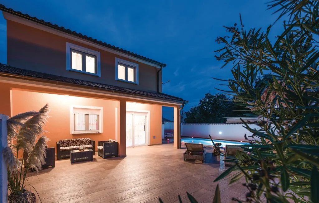 A lit house opens onto a tiled patio beside a swimming pool at dusk.