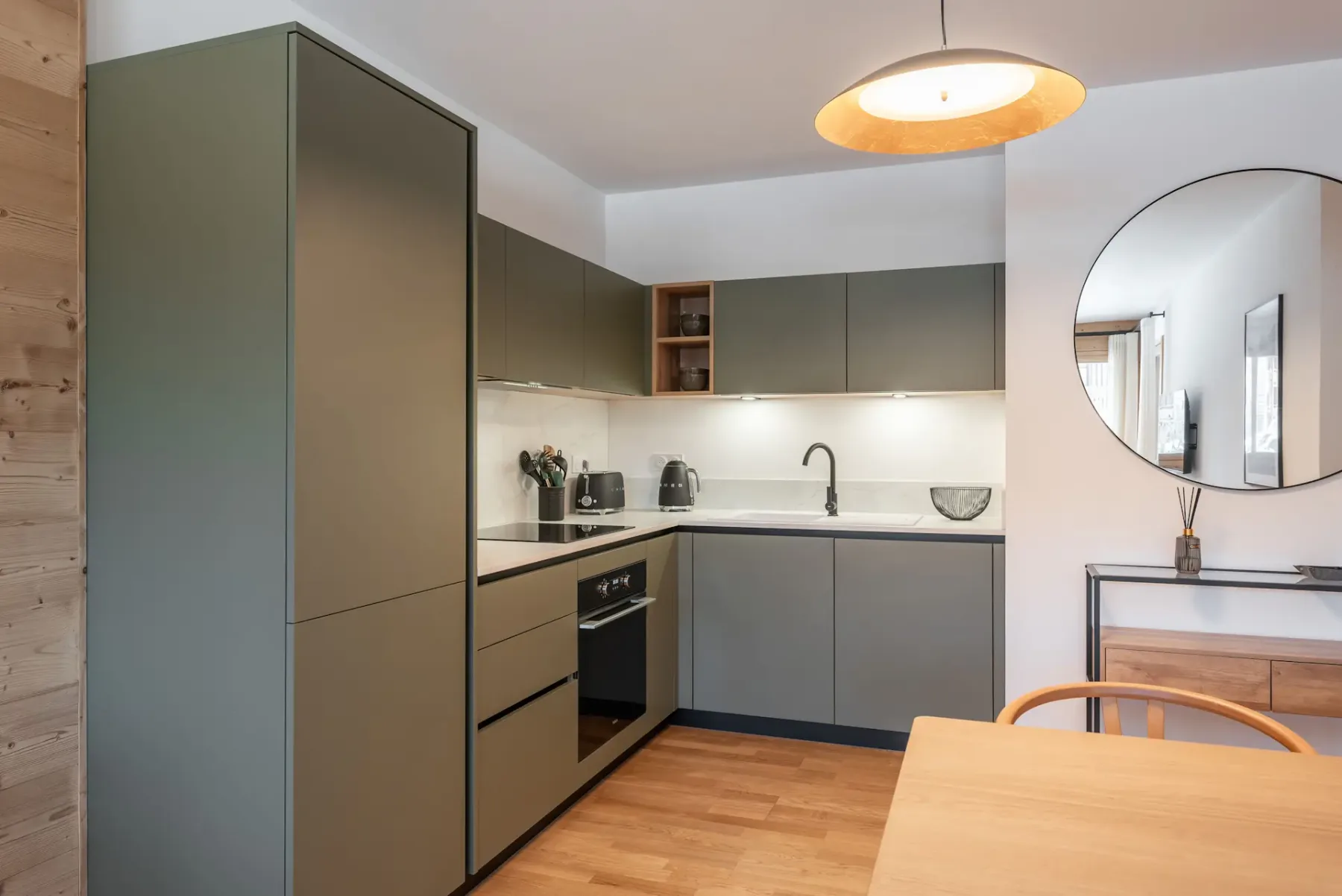 Modern kitchen with green cabinets, wooden dining table, four chairs, circular mirror, pendant light, and framed landscape artwork on the wall.