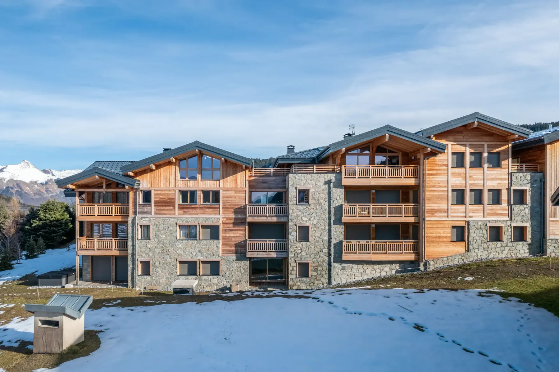 A row of modern chalet-style buildings with wood and stone exteriors sits amid a partially snow-covered landscape, surrounded by trees and other houses in a mountain village.