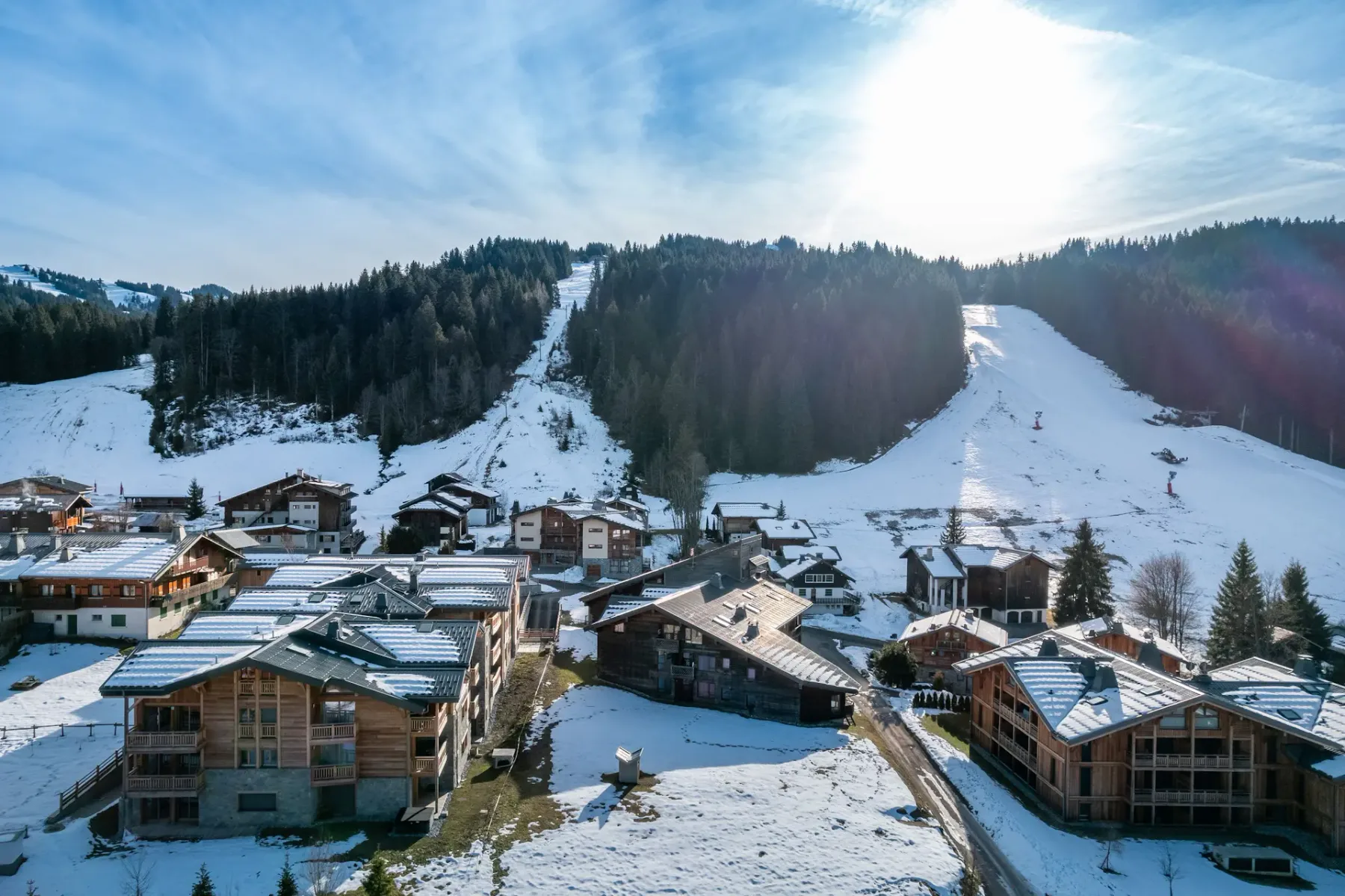 A row of modern chalet-style buildings with wood and stone exteriors sits amid a partially snow-covered landscape, surrounded by trees and other houses in a mountain village.