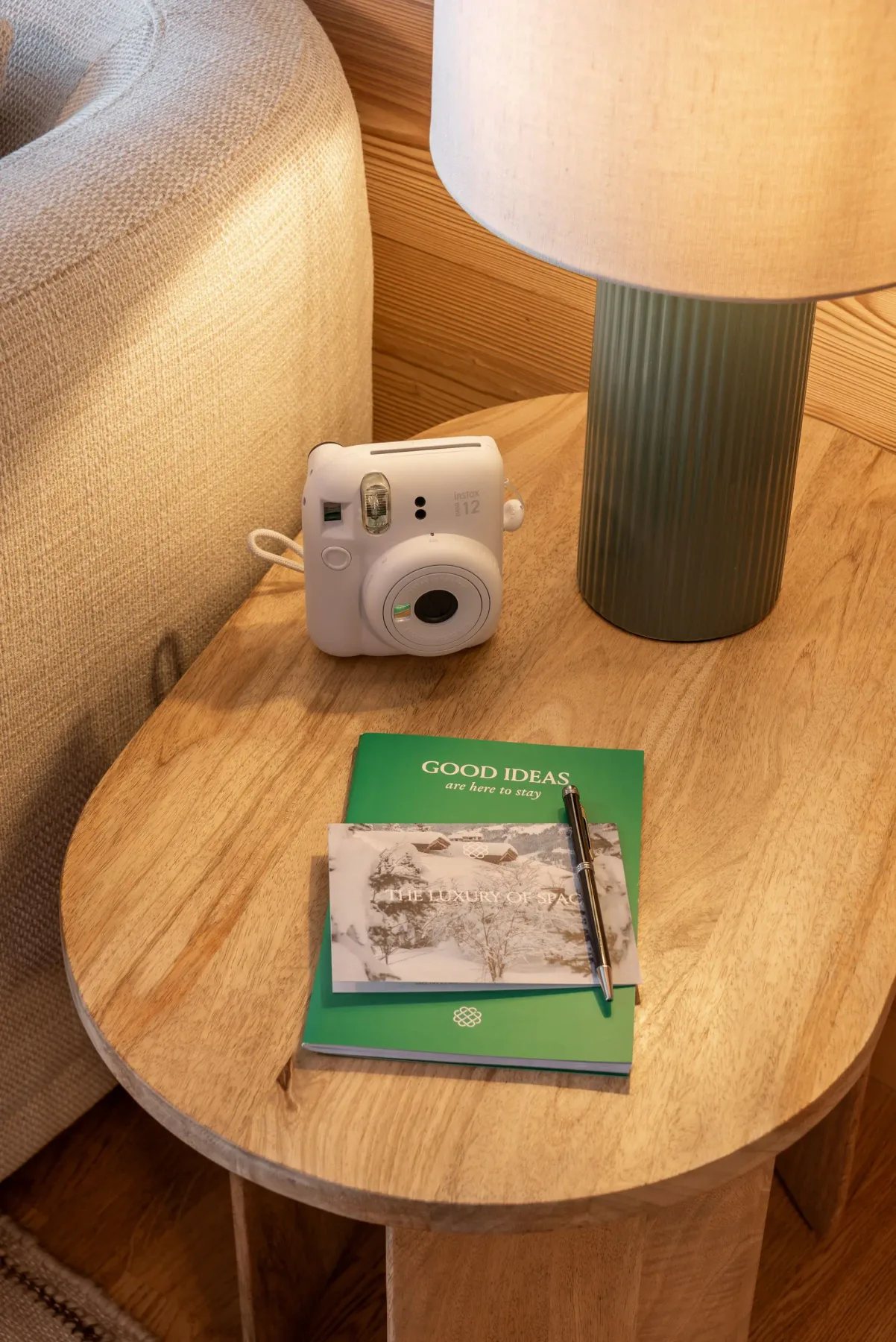 Cozy room with a beige sofa, wooden side table, lamp, instant camera, and books. Wood-paneled wall and patterned rug enhance warmth.