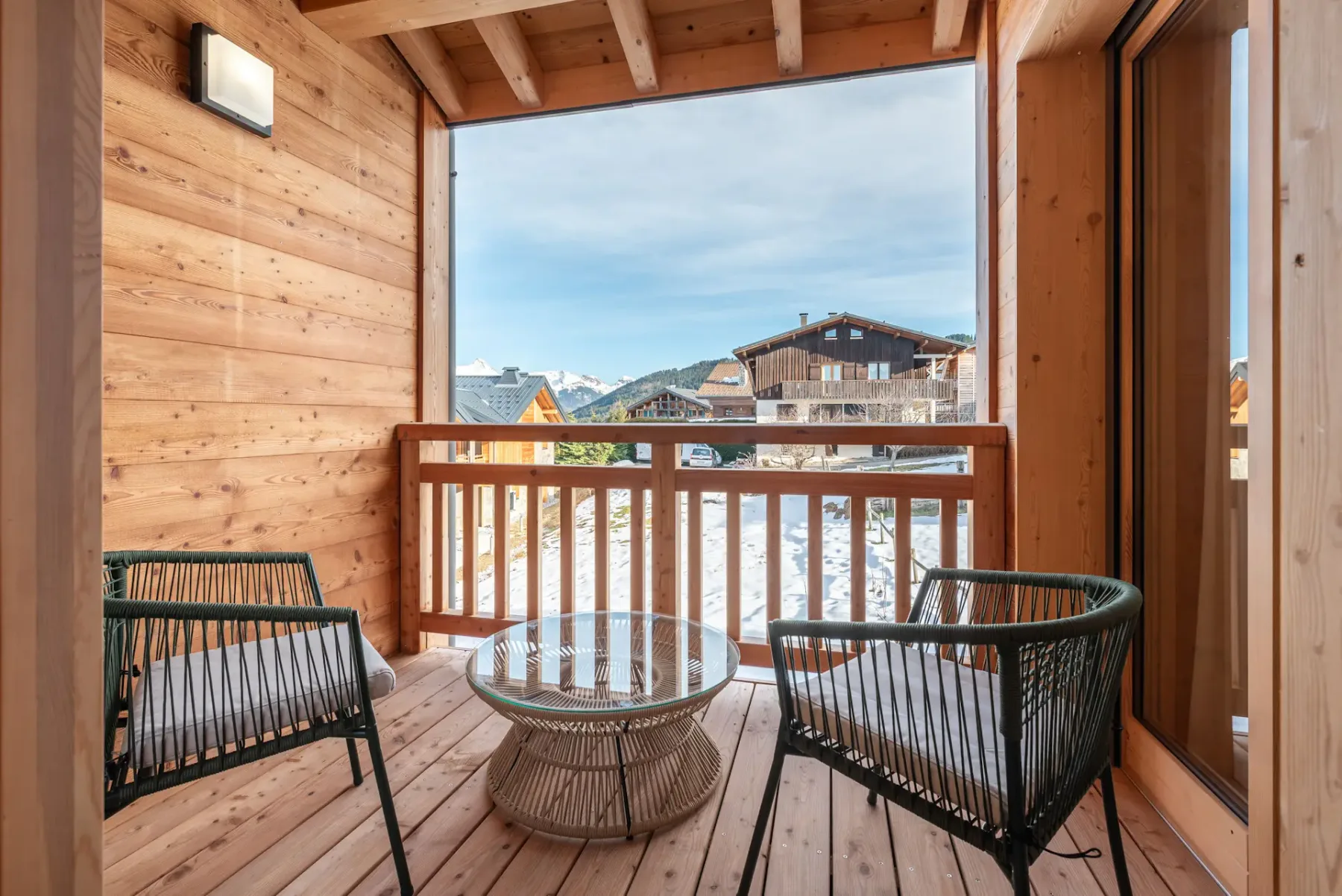 Wooden balcony with two black woven chairs and a glass-top table, overlooking snowy rooftops and a clear blue sky.
