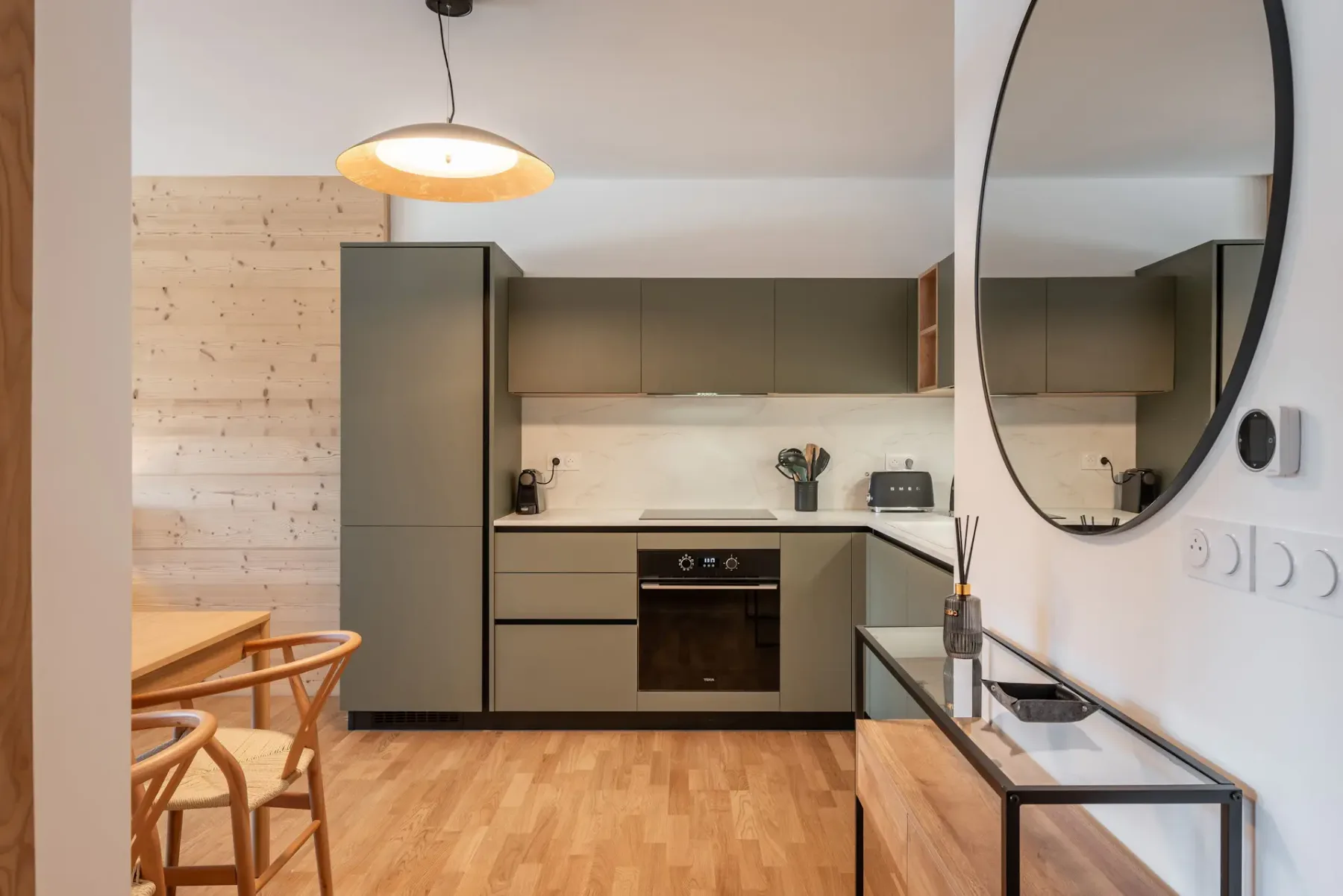 Modern kitchen with green cabinets, wooden dining table, four chairs, circular mirror, pendant light, and framed landscape artwork on the wall.
