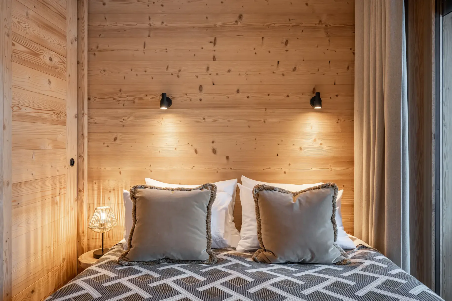 Cozy bedroom with wooden walls, a double bed, and a geometric bedspread. Glass doors open to a balcony with mountain views.