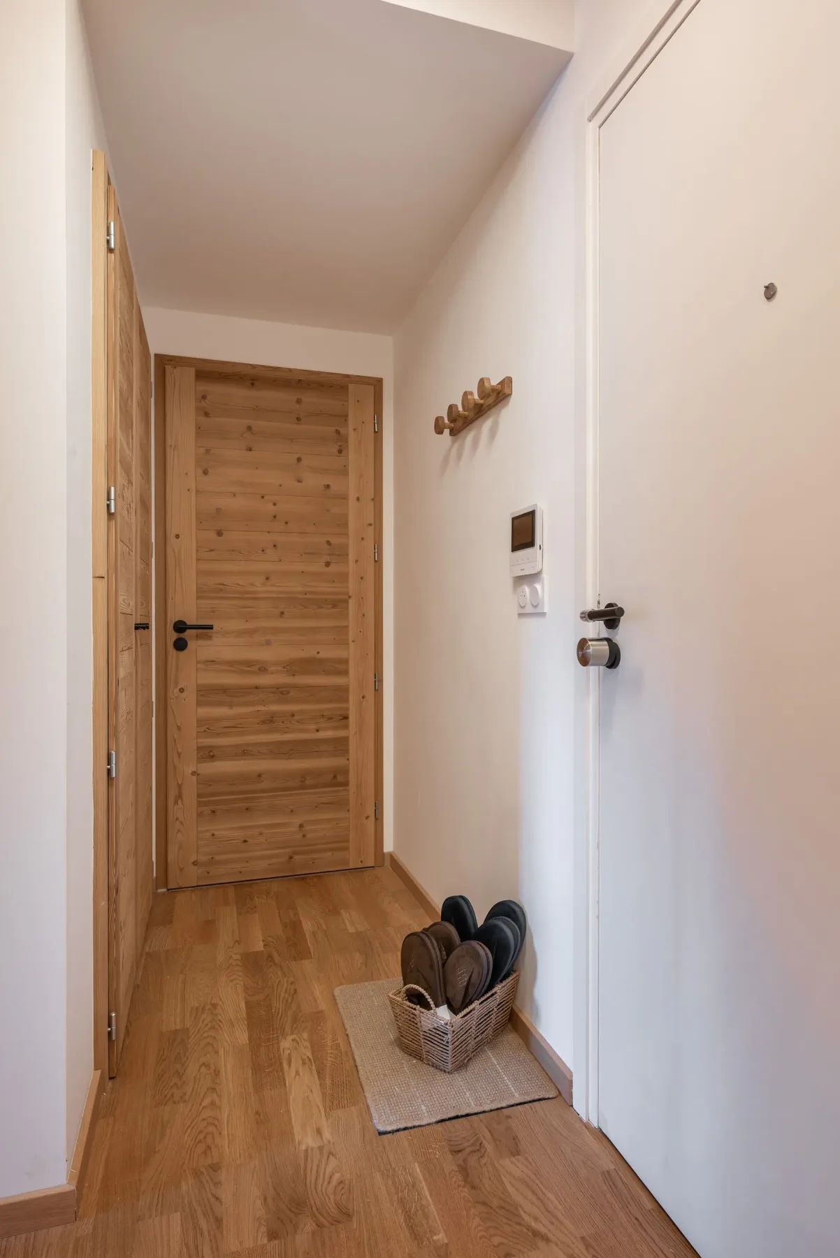 Hallway with wooden flooring, white walls, a wooden door, coat hooks, light switch, and a basket of shoes by the door on a small rug.