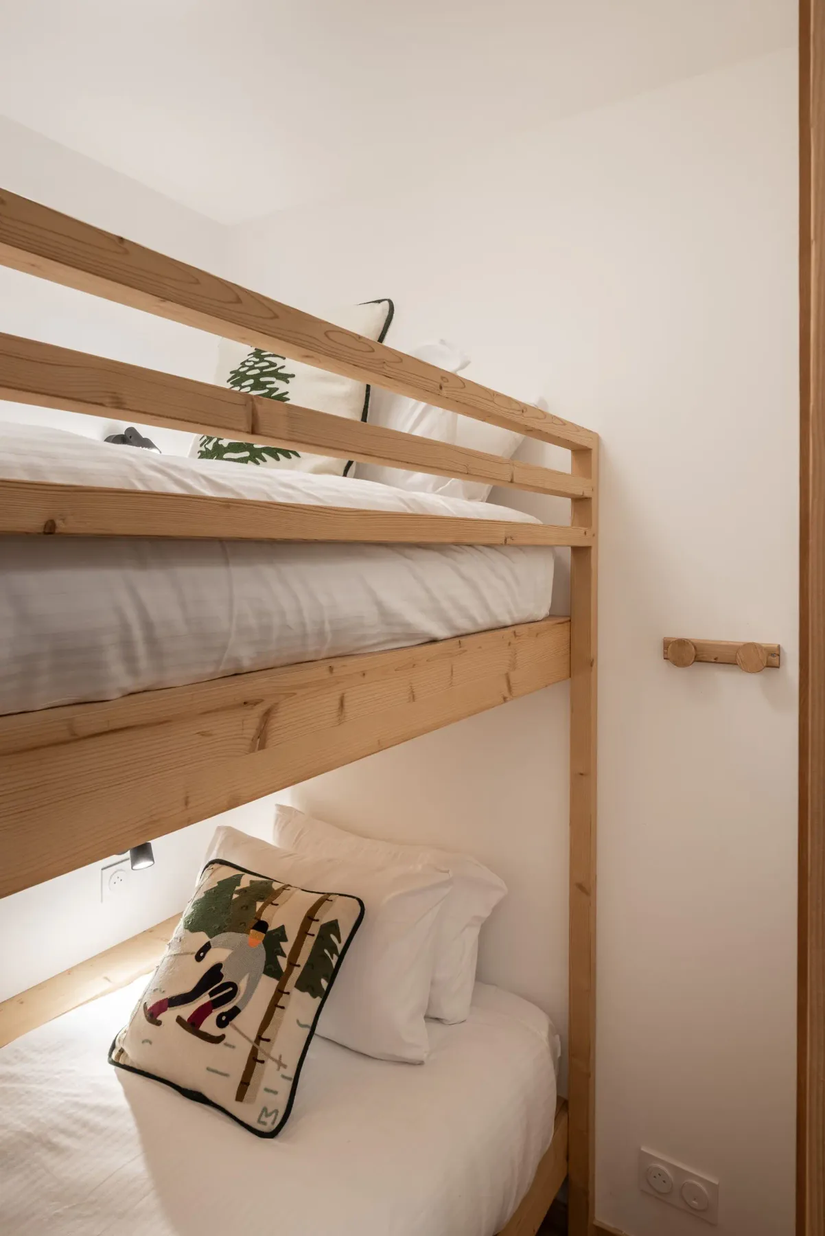 Wooden bunk bed with white bedding in a minimalistic, well-lit room. A decorative pillow and small plant add a touch of coziness.