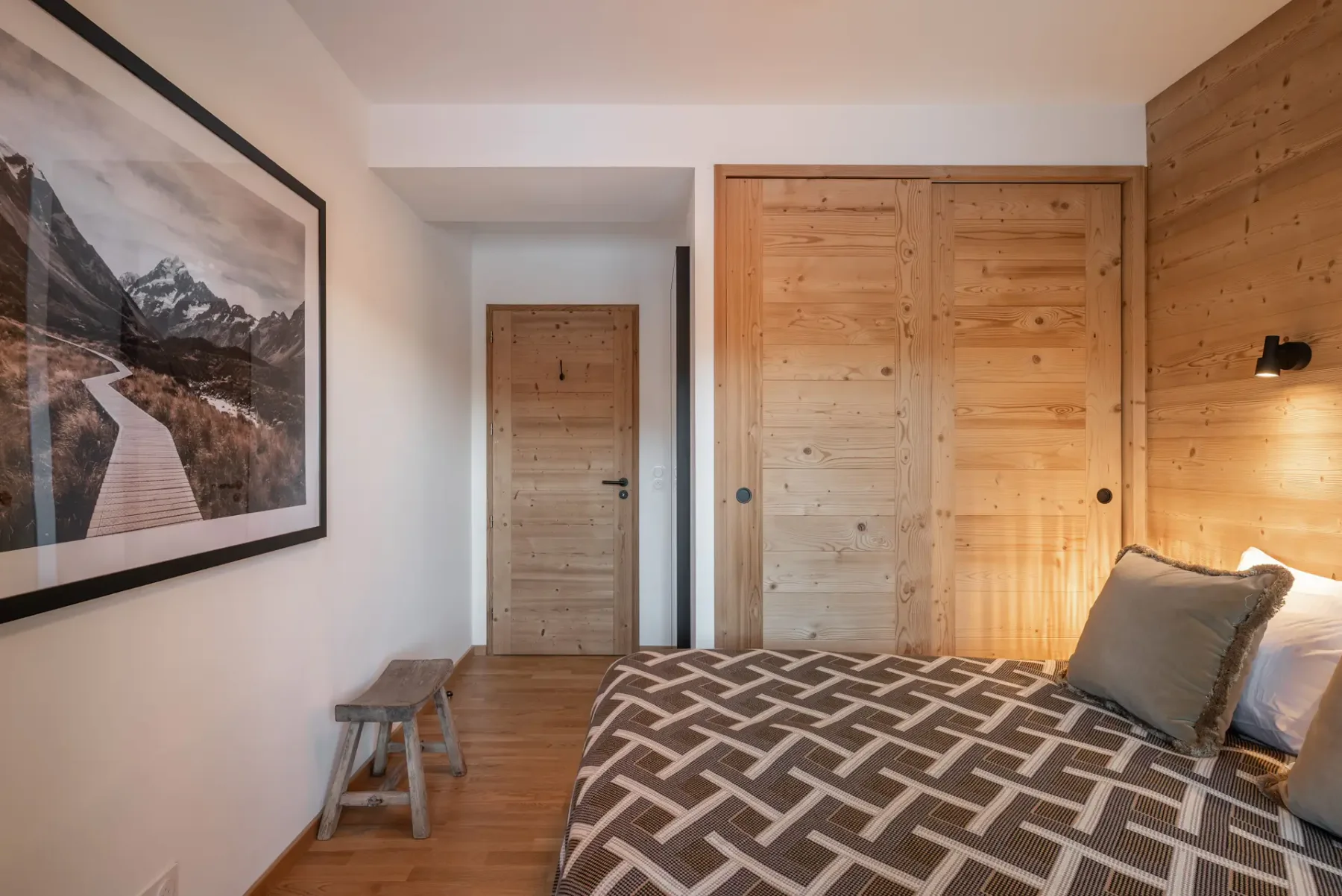 Cozy bedroom with wooden walls, a double bed, and a geometric bedspread. Glass doors open to a balcony with mountain views.