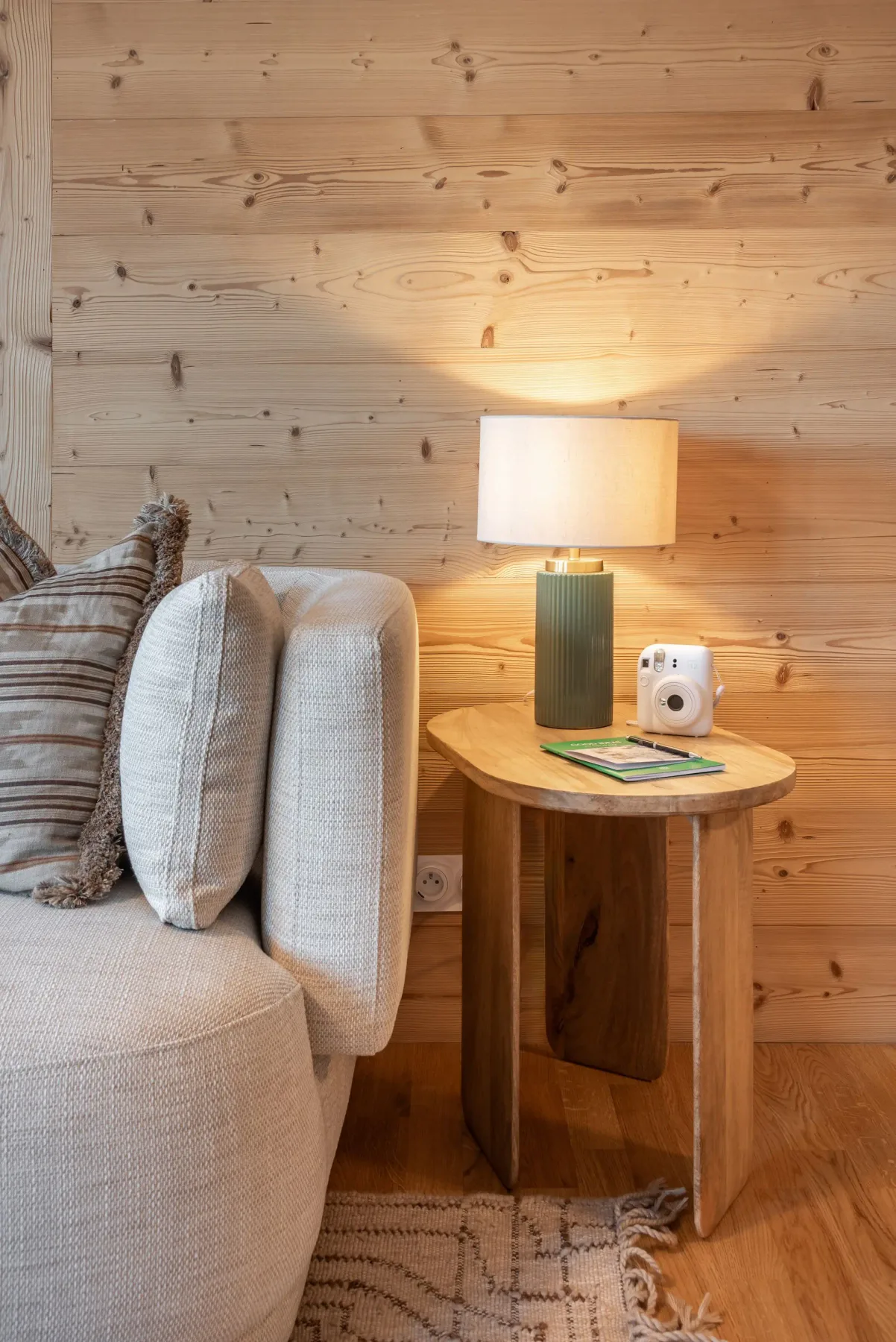 Cozy room with a beige sofa, wooden side table, lamp, instant camera, and books. Wood-paneled wall and patterned rug enhance warmth.