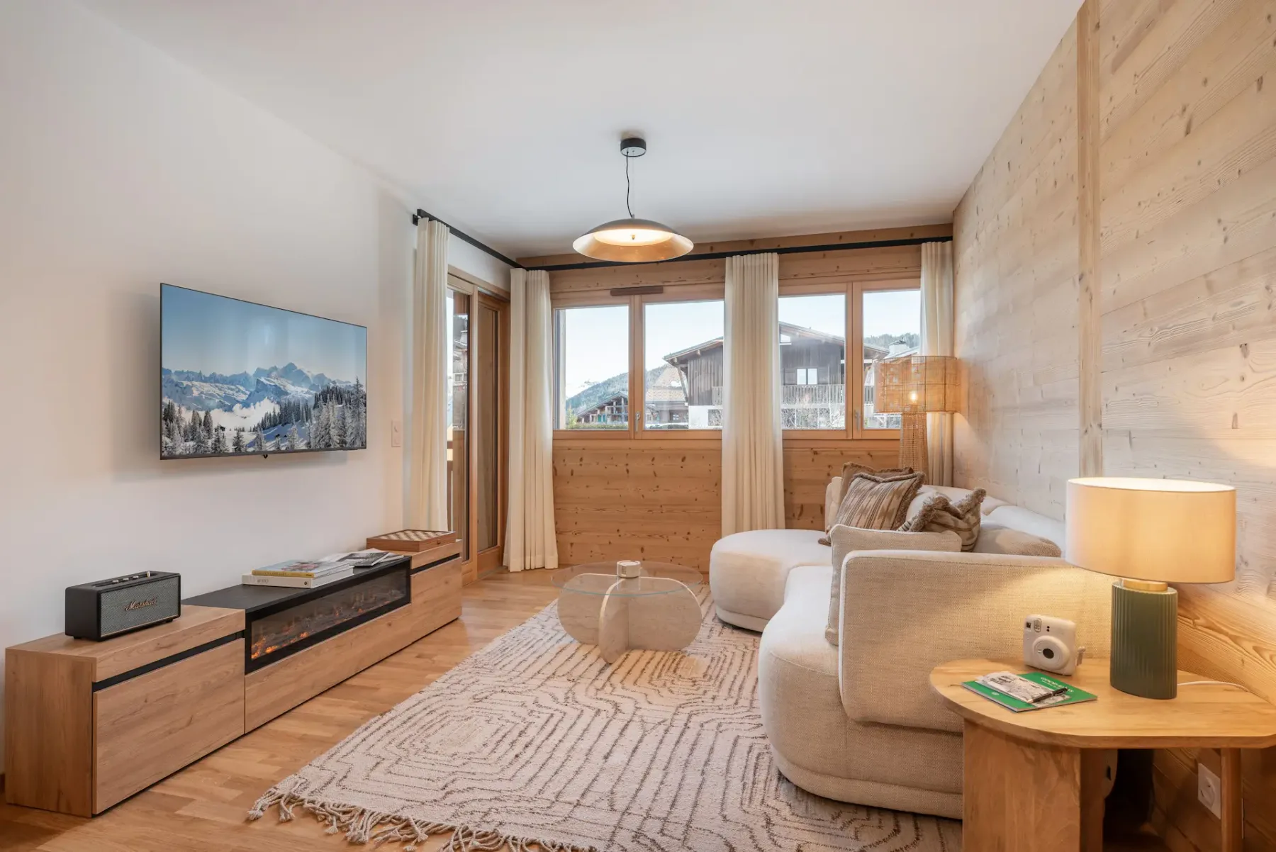 Cozy living room with a beige curved sofa, wooden walls, large windows, a patterned rug, and modern lamp. Light and airy space.