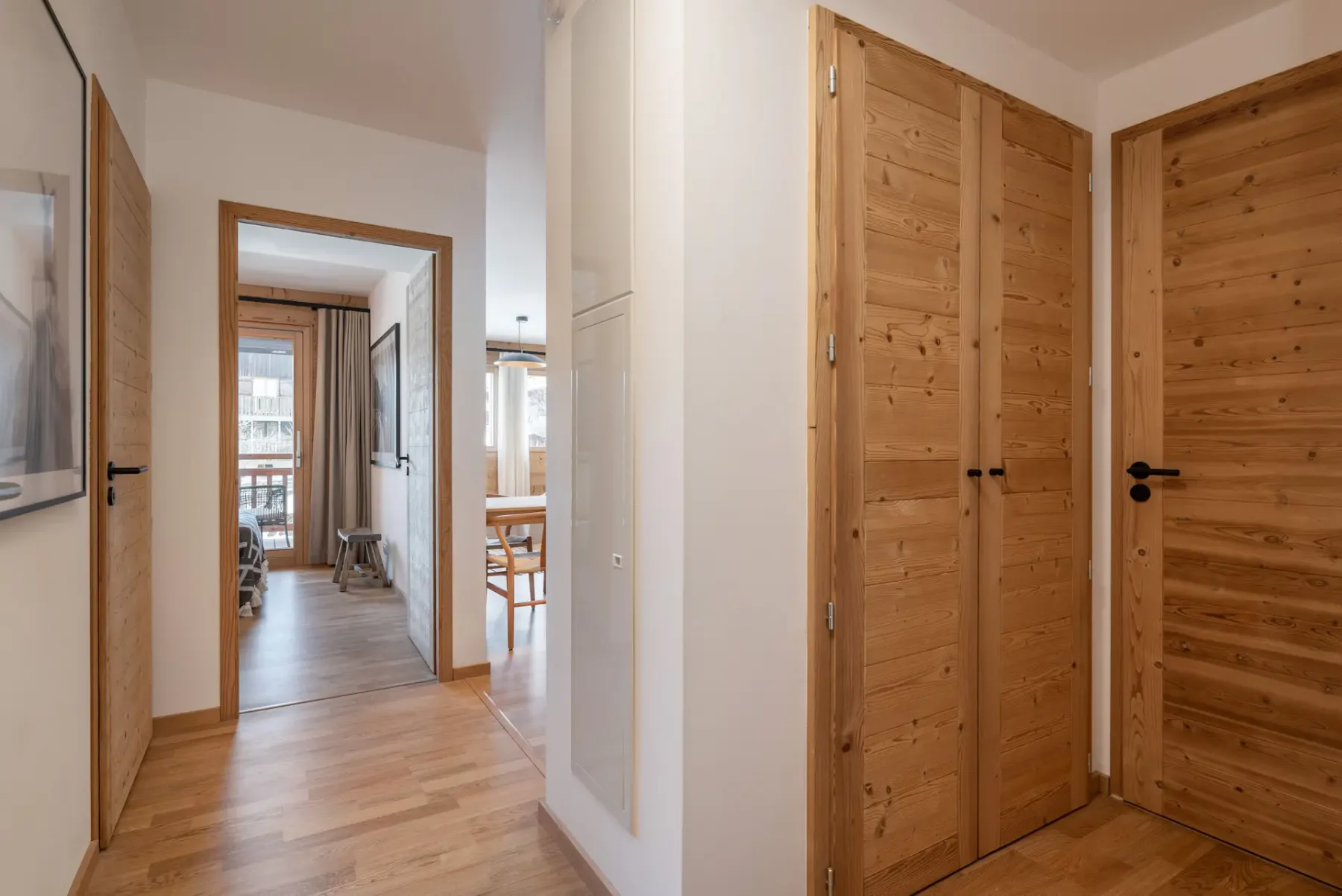 A hallway with light wooden flooring, two wooden doors on the right, and an open door leading to a bright room with a table and chairs.