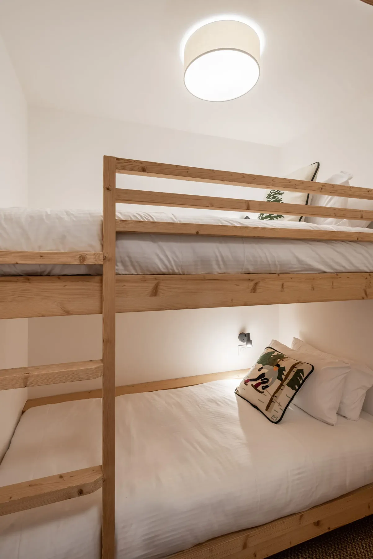 Wooden bunk bed with white bedding in a minimalistic, well-lit room. A decorative pillow and small plant add a touch of coziness.
