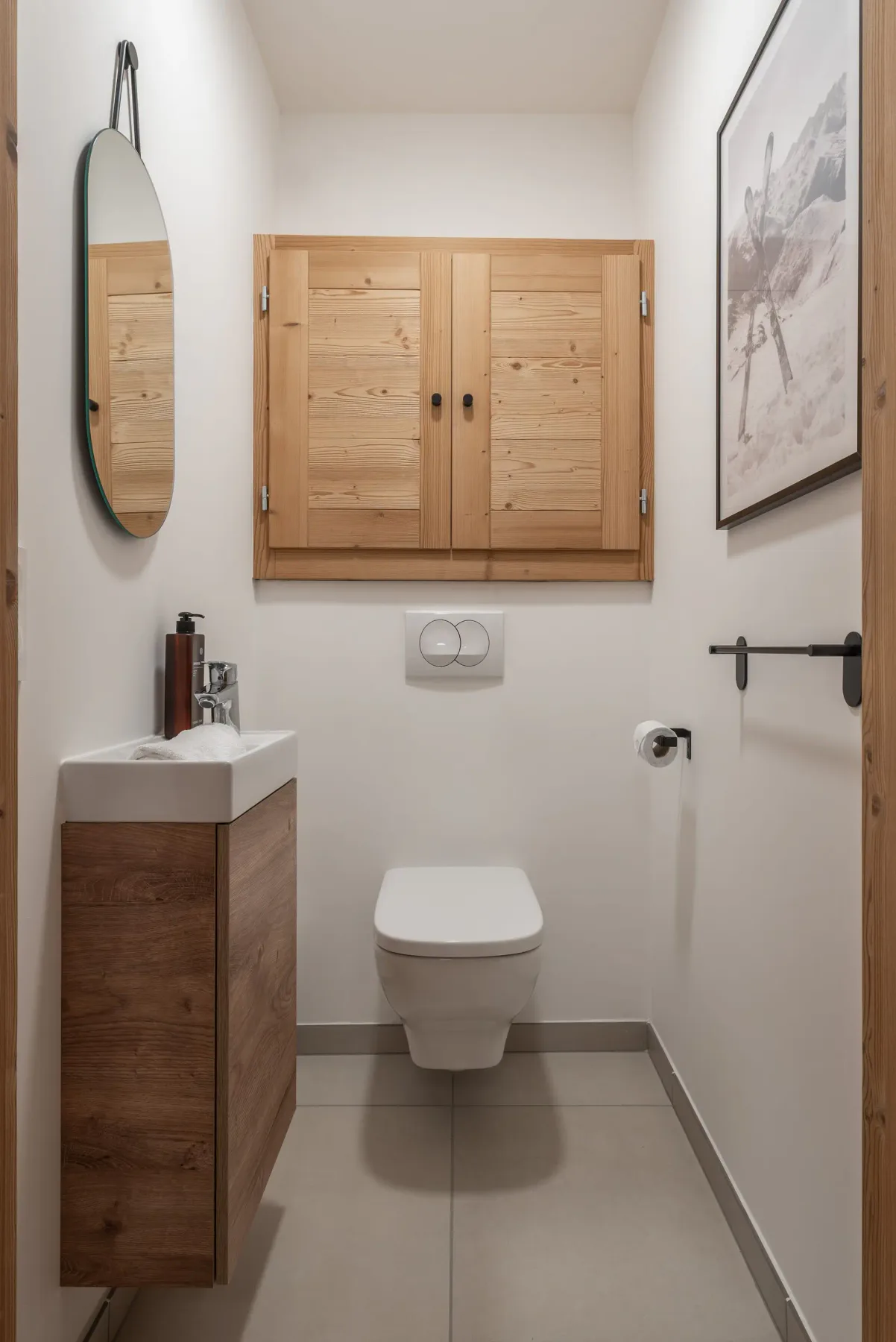 A small bathroom with white walls features a black sink, wooden accents, a round mirror, and a toilet. The toilet paper holder is mounted on the wall next to the sink. A decorative piece rests on a wooden shelf.