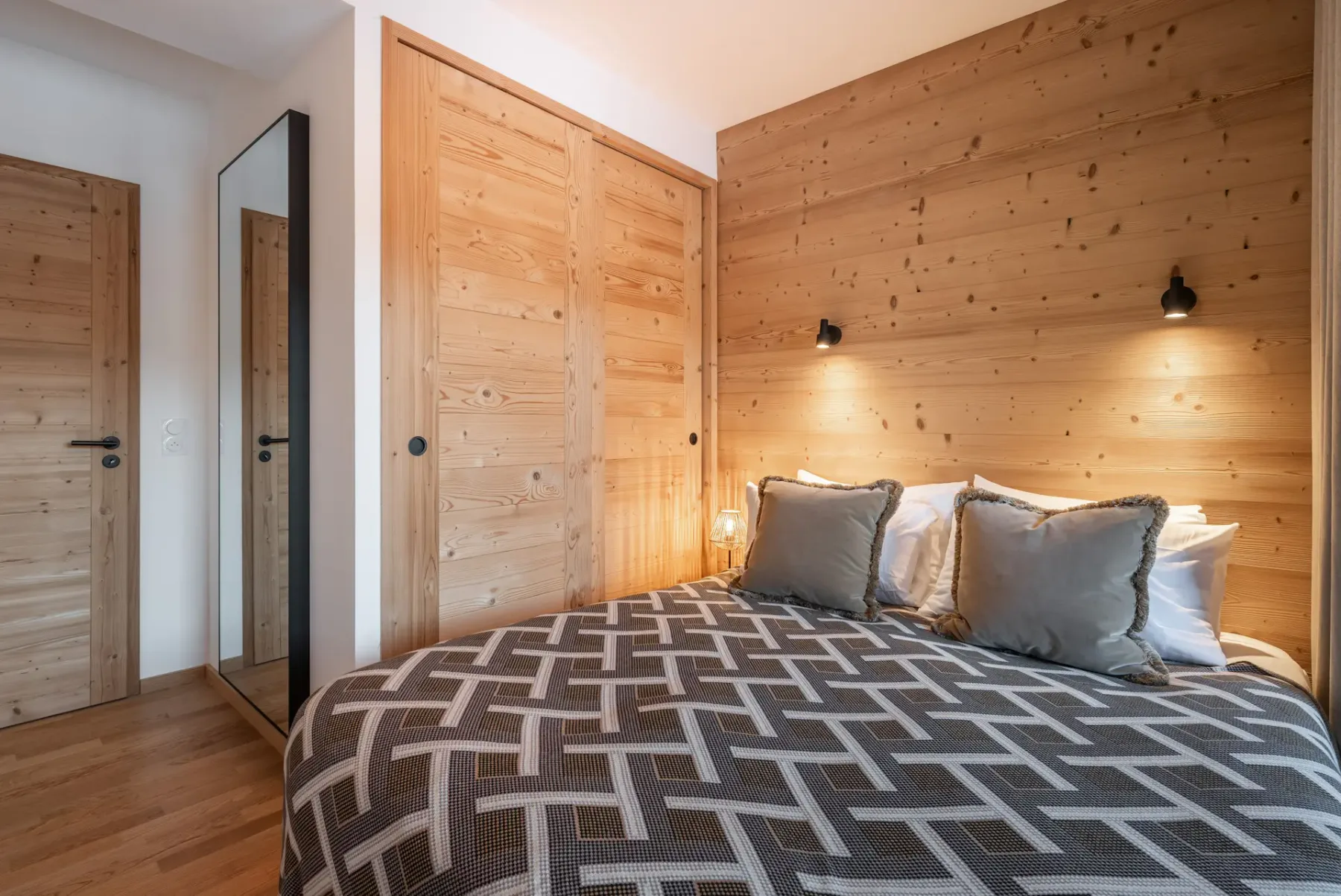 Cozy bedroom with wooden walls, a double bed, and a geometric bedspread. Glass doors open to a balcony with mountain views.