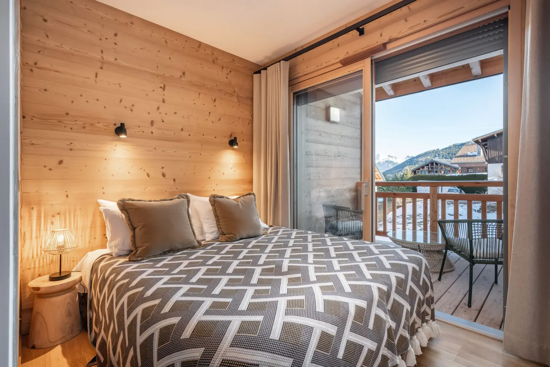 Cozy bedroom with wooden walls, a double bed, and a geometric bedspread. Glass doors open to a balcony with mountain views.