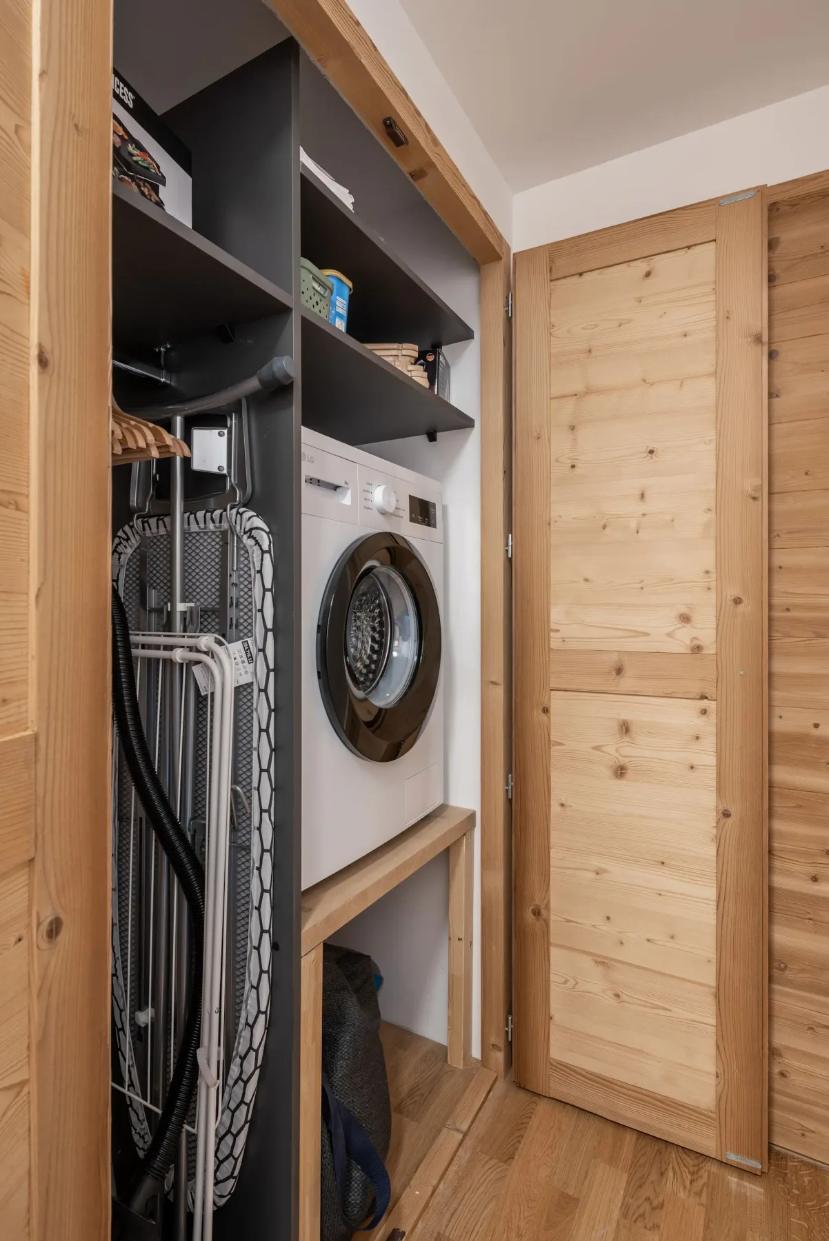 Compact laundry space with a front-loading washing machine, ironing board, and shelves, enclosed by wooden panels.