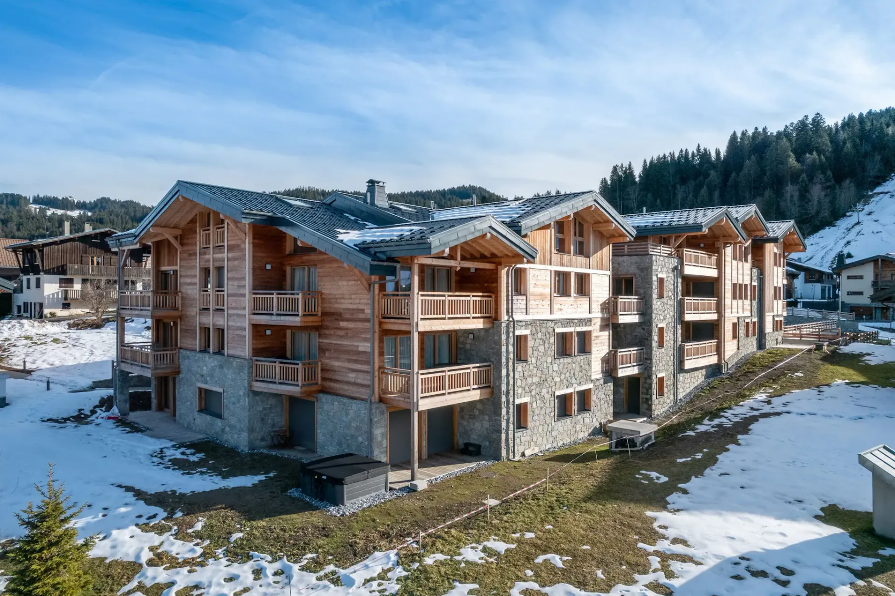A row of modern chalet-style buildings with wood and stone exteriors sits amid a partially snow-covered landscape, surrounded by trees and other houses in a mountain village.