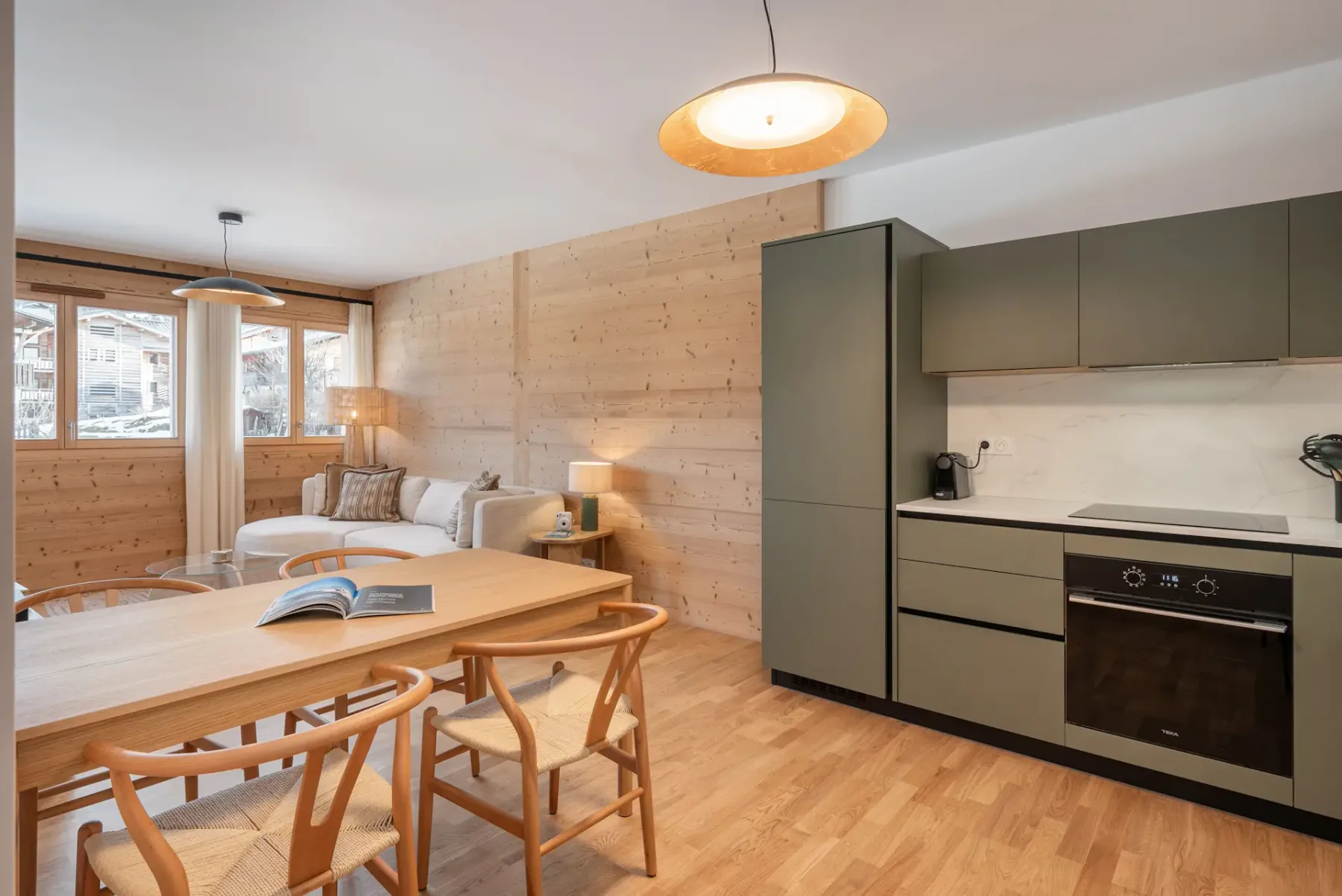 Modern kitchen with green cabinets, wooden dining table, four chairs, circular mirror, pendant light, and framed landscape artwork on the wall.