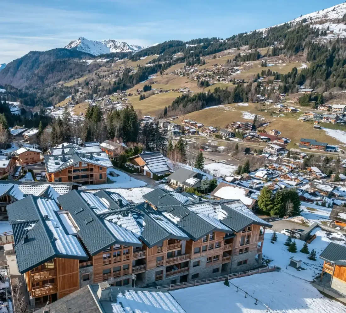 A row of modern chalet-style buildings with wood and stone exteriors sits amid a partially snow-covered landscape, surrounded by trees and other houses in a mountain village.