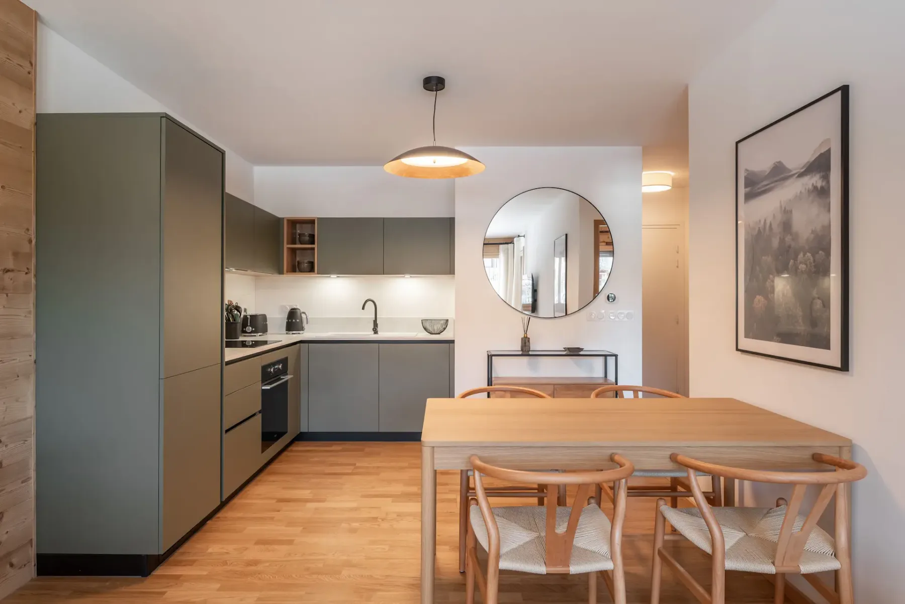 Modern kitchen with green cabinets, wooden dining table, four chairs, circular mirror, pendant light, and framed landscape artwork on the wall.