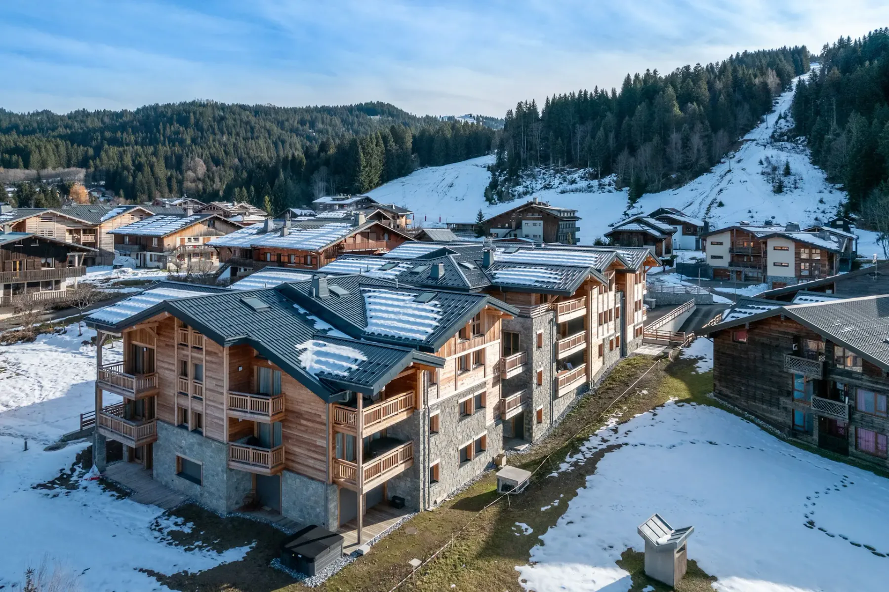 A row of modern chalet-style buildings with wood and stone exteriors sits amid a partially snow-covered landscape, surrounded by trees and other houses in a mountain village.