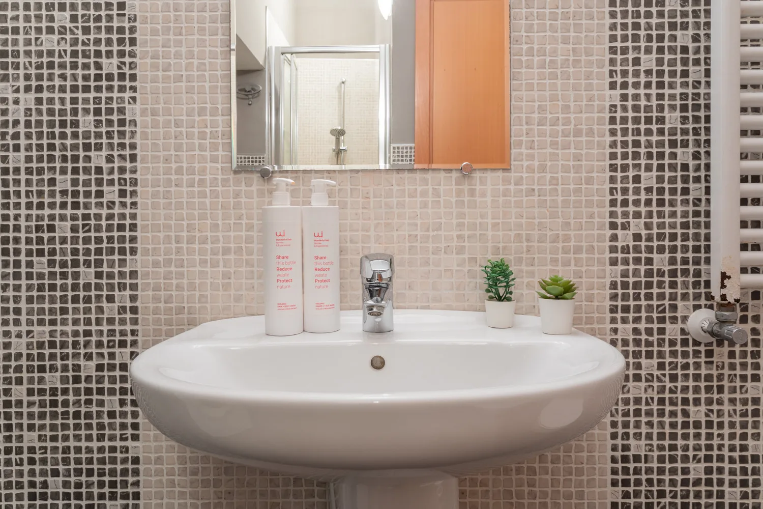 Un lavabo de baño está bajo un espejo, con paredes de azulejos y pequeñas plantas.