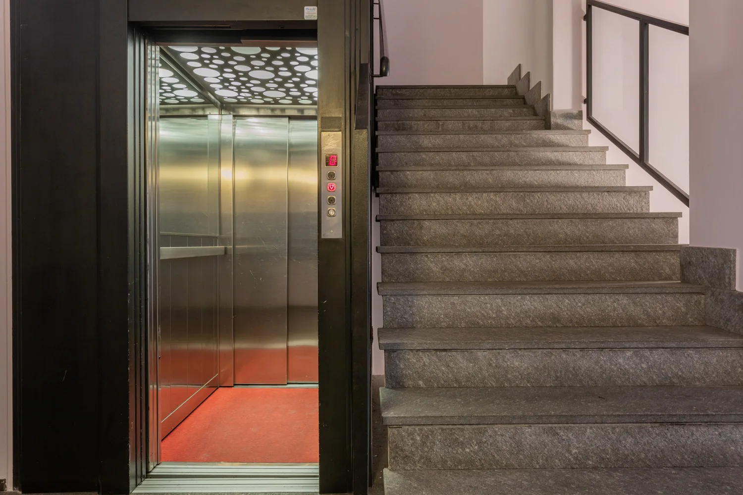 Un ascensor abierto está junto a una escalera con pasamanos.