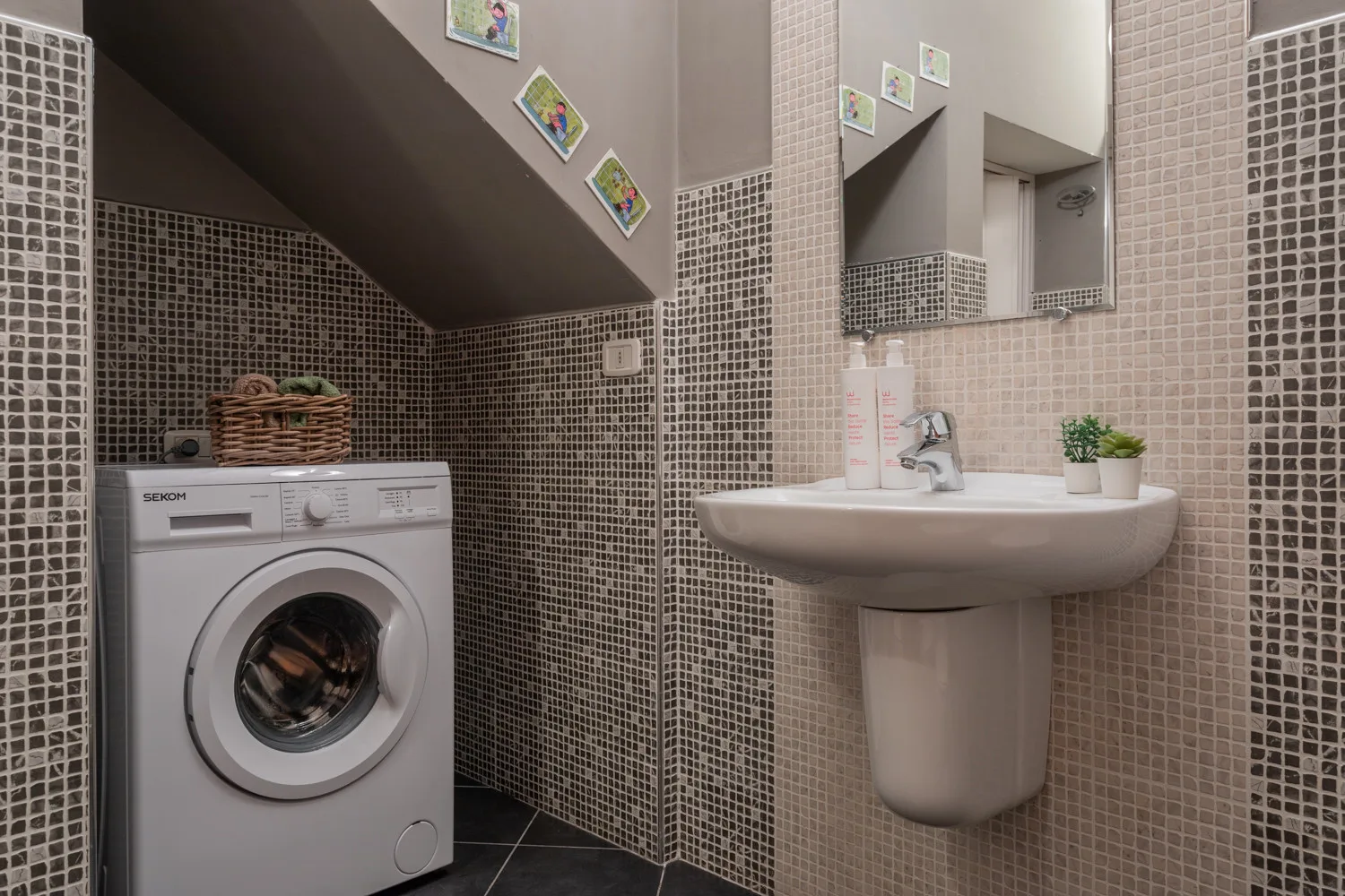 Un baño alicatado con lavabo de pedestal y lavadora bajo un techo inclinado.