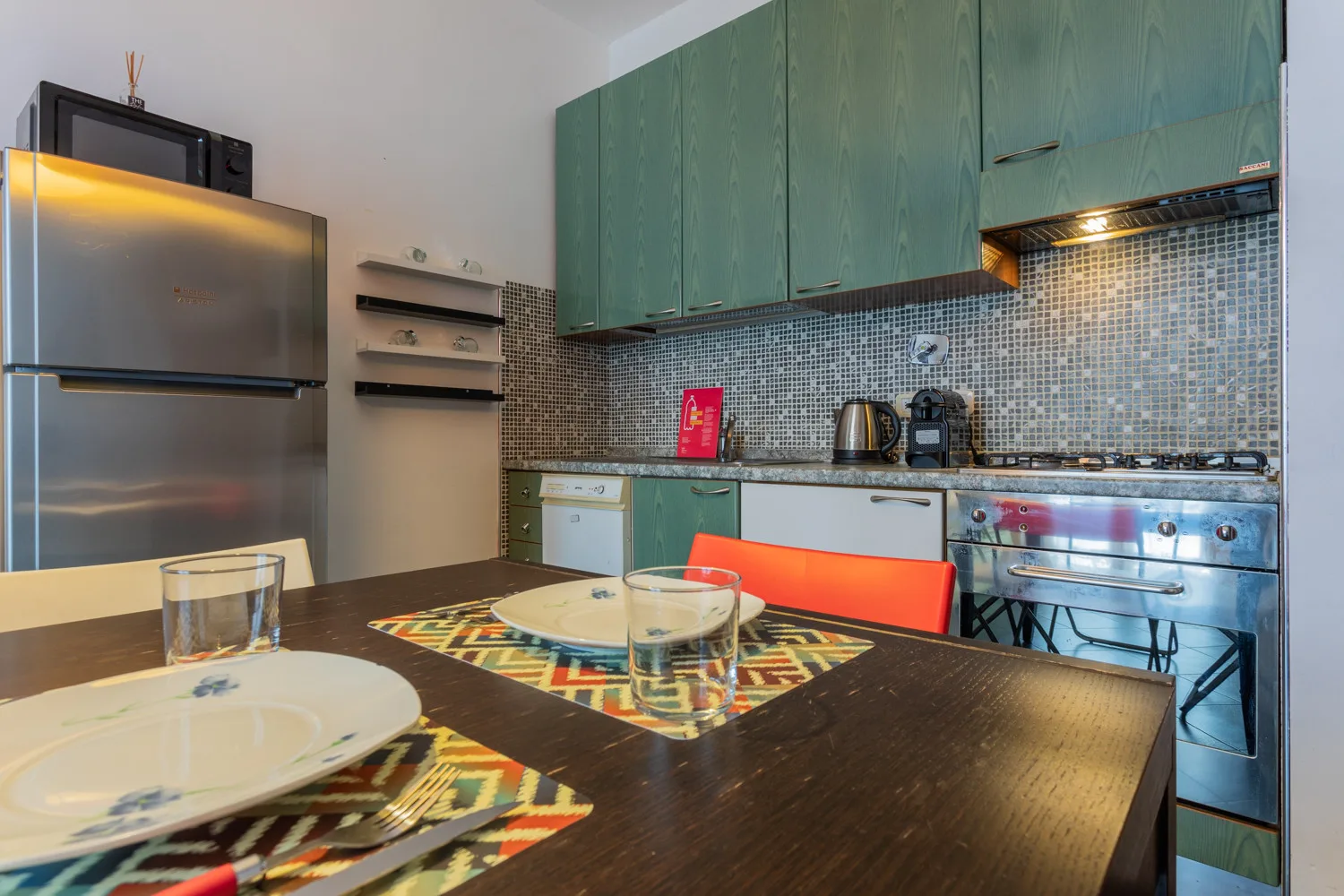 Una mesa de comedor está frente a una cocina con gabinetes verdes y electrodomésticos de acero inoxidable.